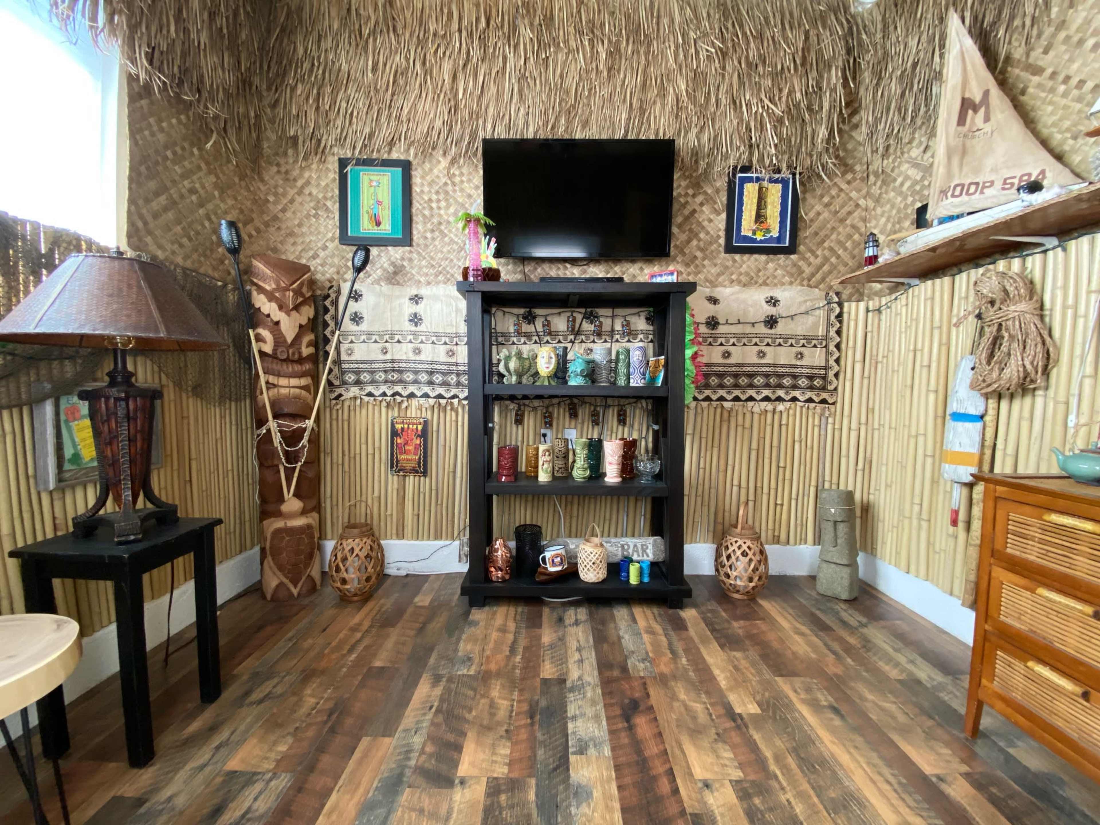 The room features a thatched wall design, a black shelf filled with colorful cups, a television mounted above it, and various decorative items scattered throughout.