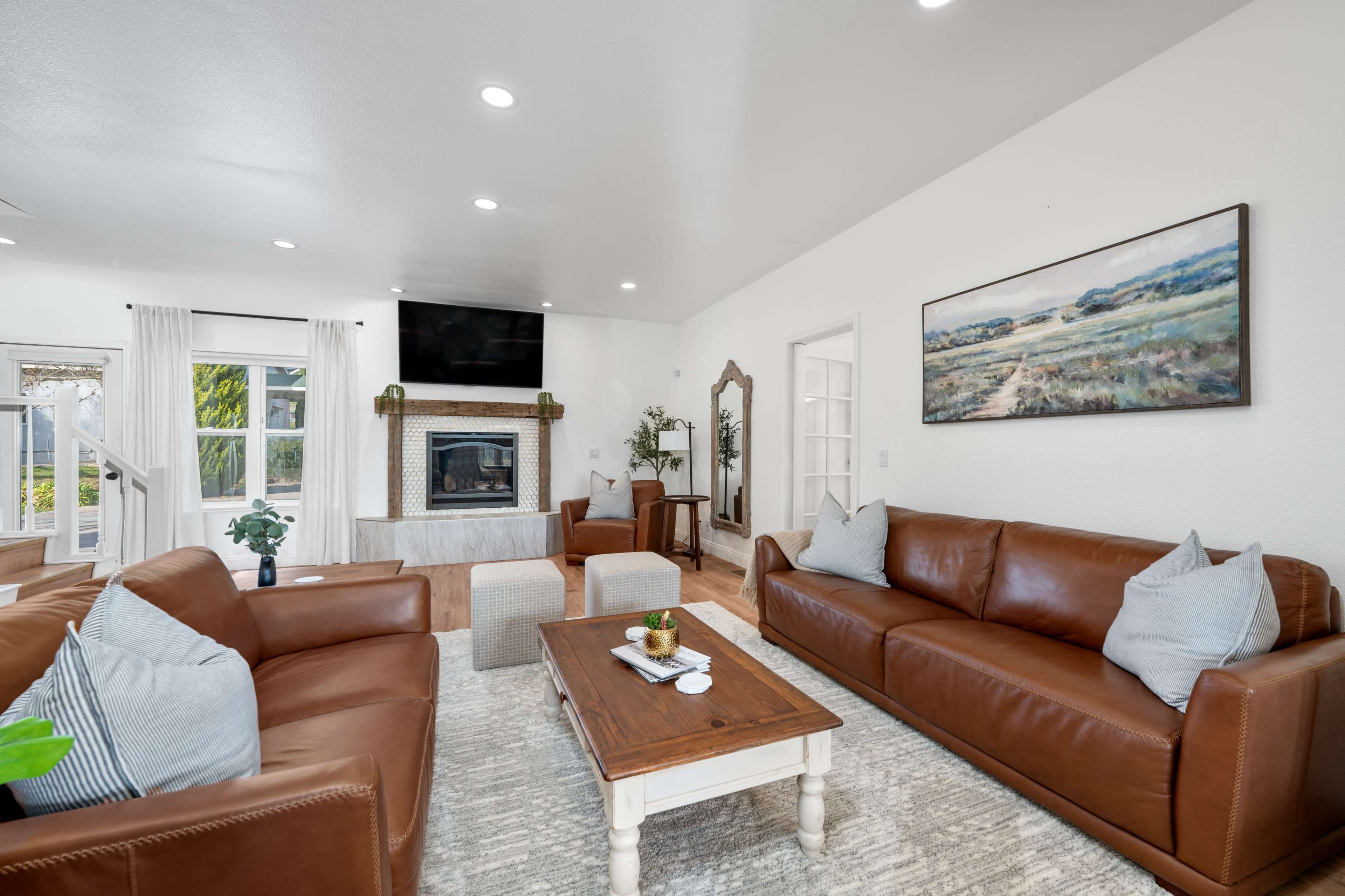 The living room features two brown leather sofas, a wooden coffee table, a fireplace with a TV above it, and large windows allowing natural light to fill the space.