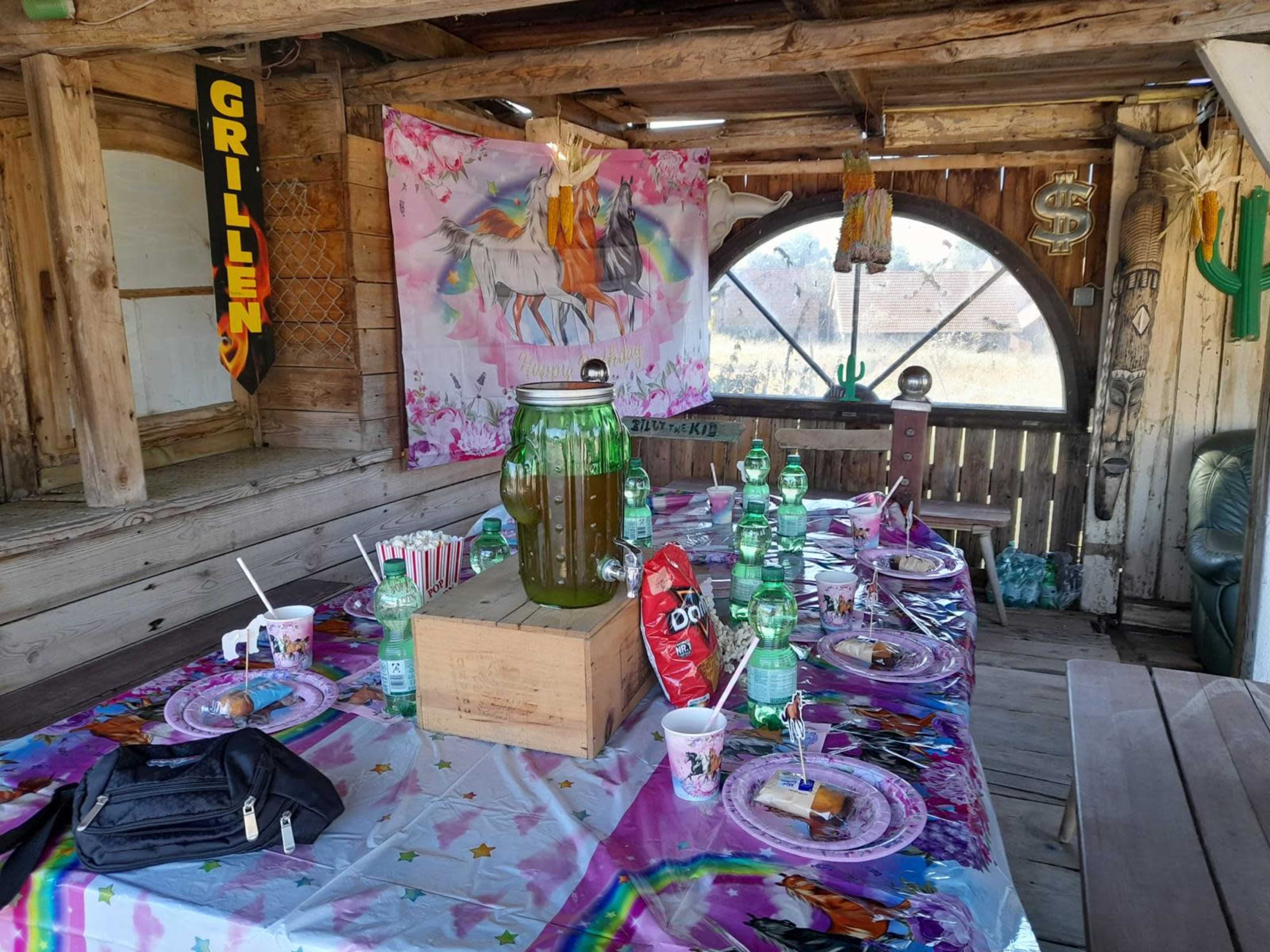 A wooden table is set for a party with colorful plates, cups, and a large jar of green drink, surrounded by decorations including a unicorn-themed backdrop.