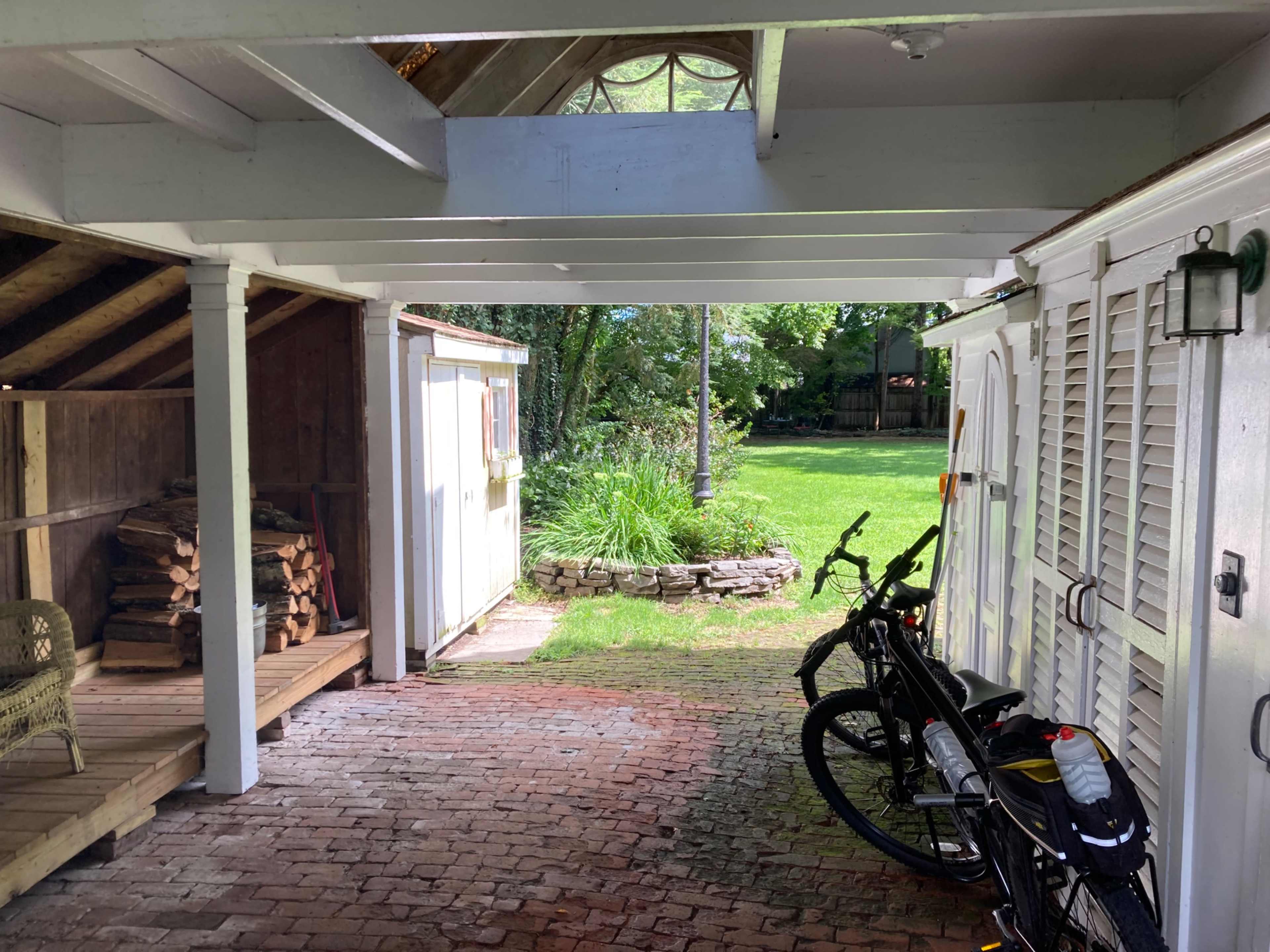 A covered, brick-paved area with bicycles parked beside a wooden storage shed and a view of a grassy yard beyond.
