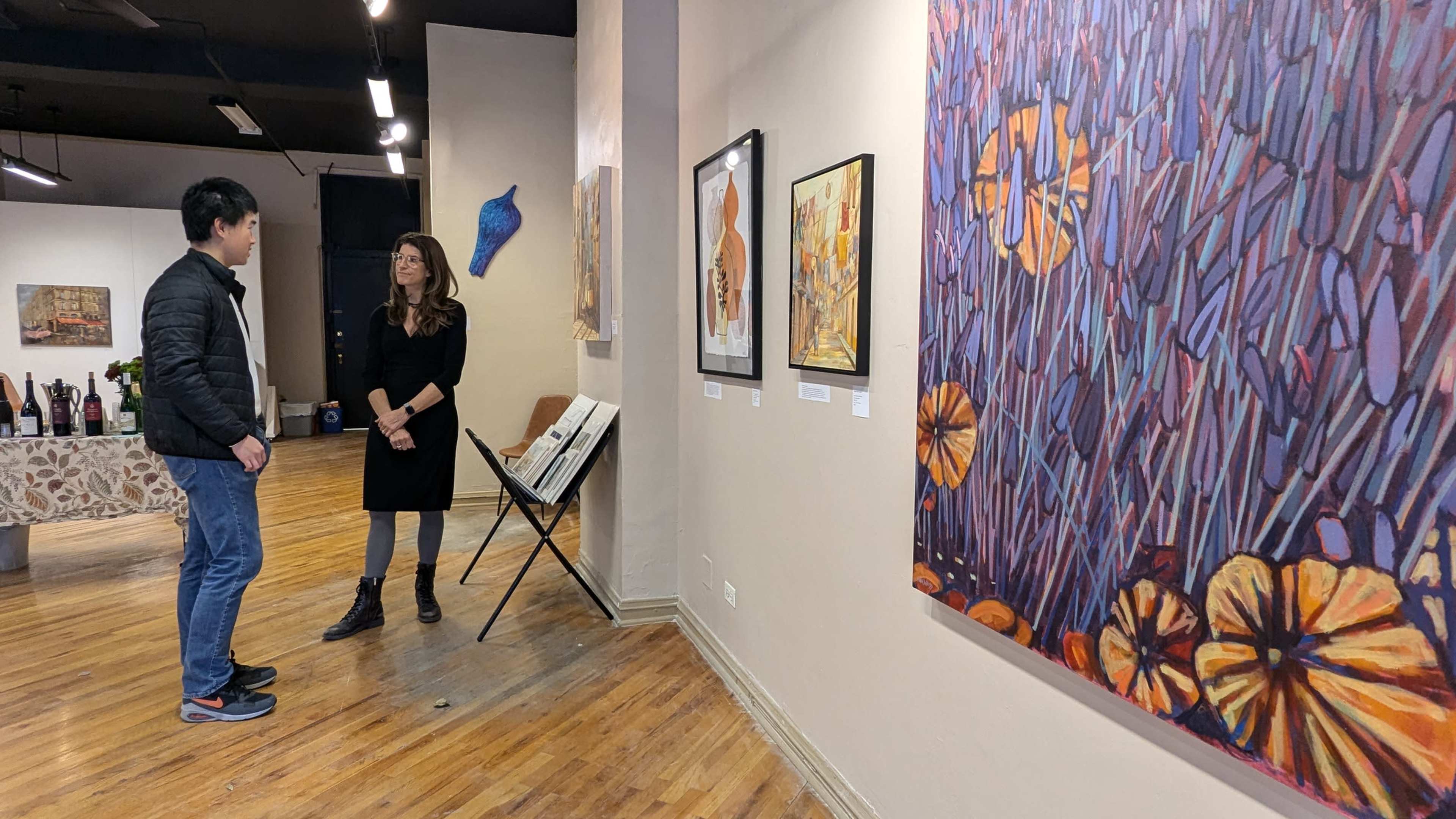 A man and a woman are conversing in an art gallery, surrounded by colorful artwork on the walls and a display table in the background.
