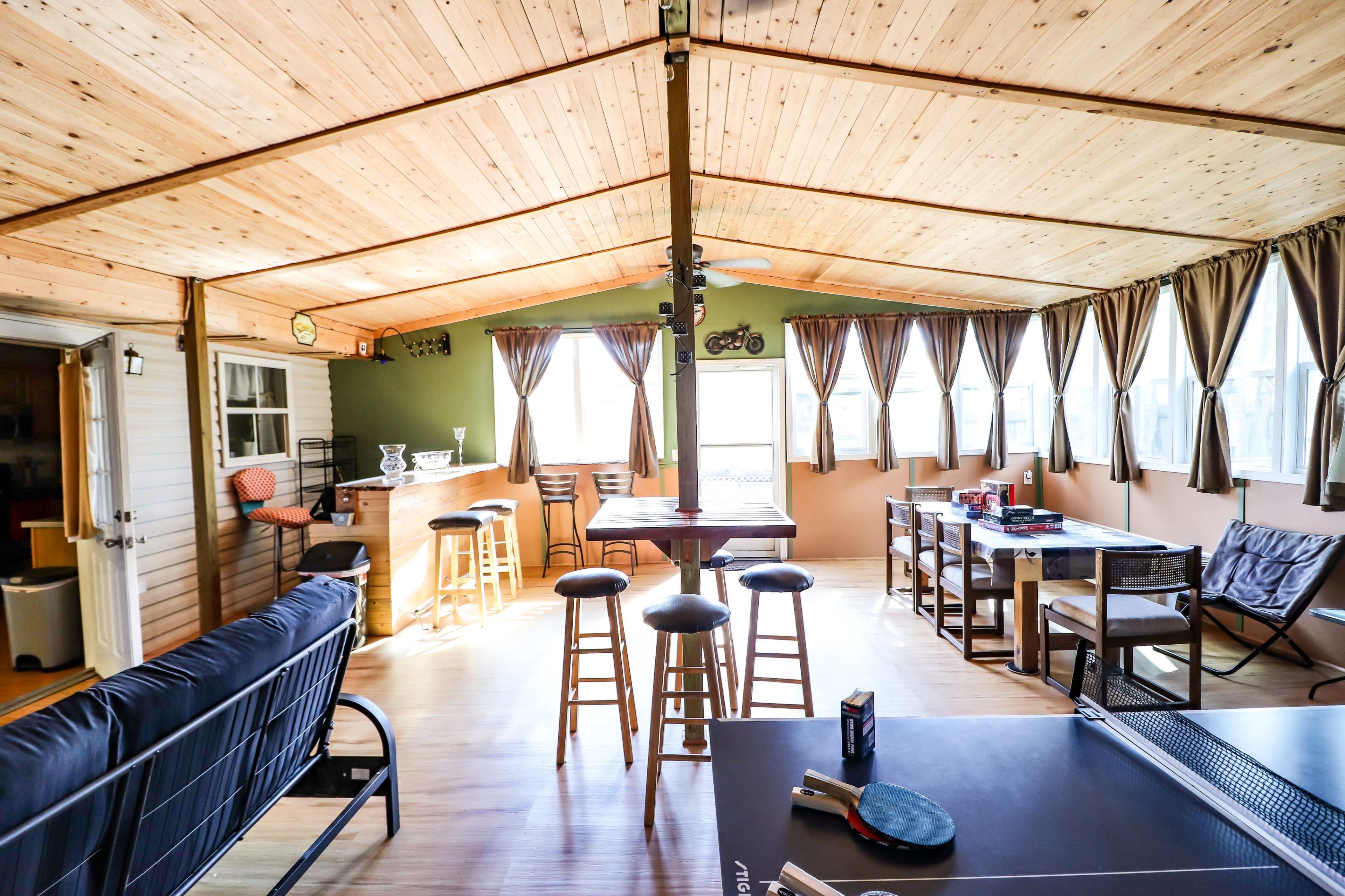The image depicts a spacious indoor sunroom with a wooden ceiling, a bar area, seating, and large windows allowing natural light.