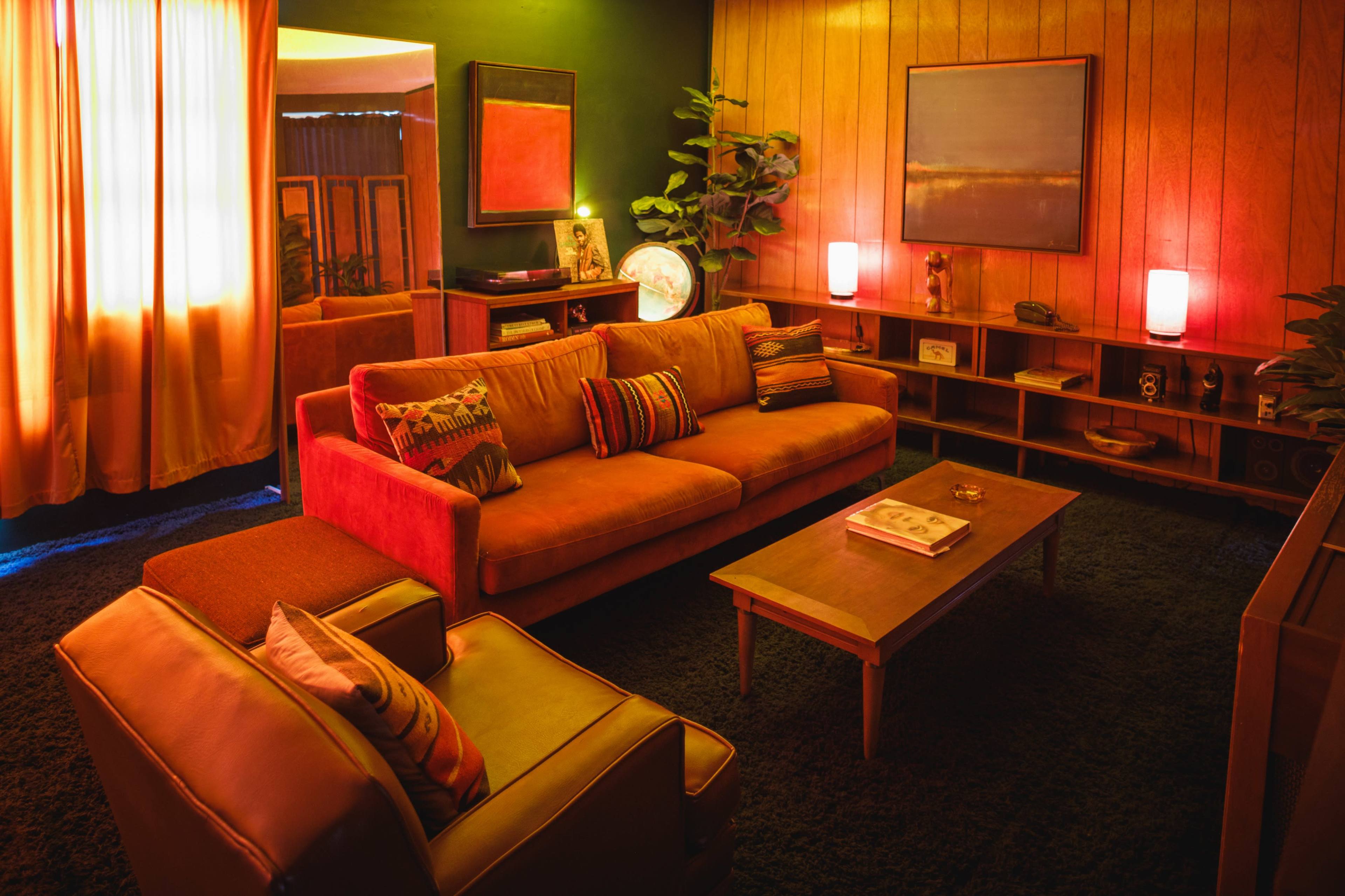 A cozy living room features a large orange sofa, a wooden coffee table, and decorative plants against wooden paneling and vibrant curtains.
