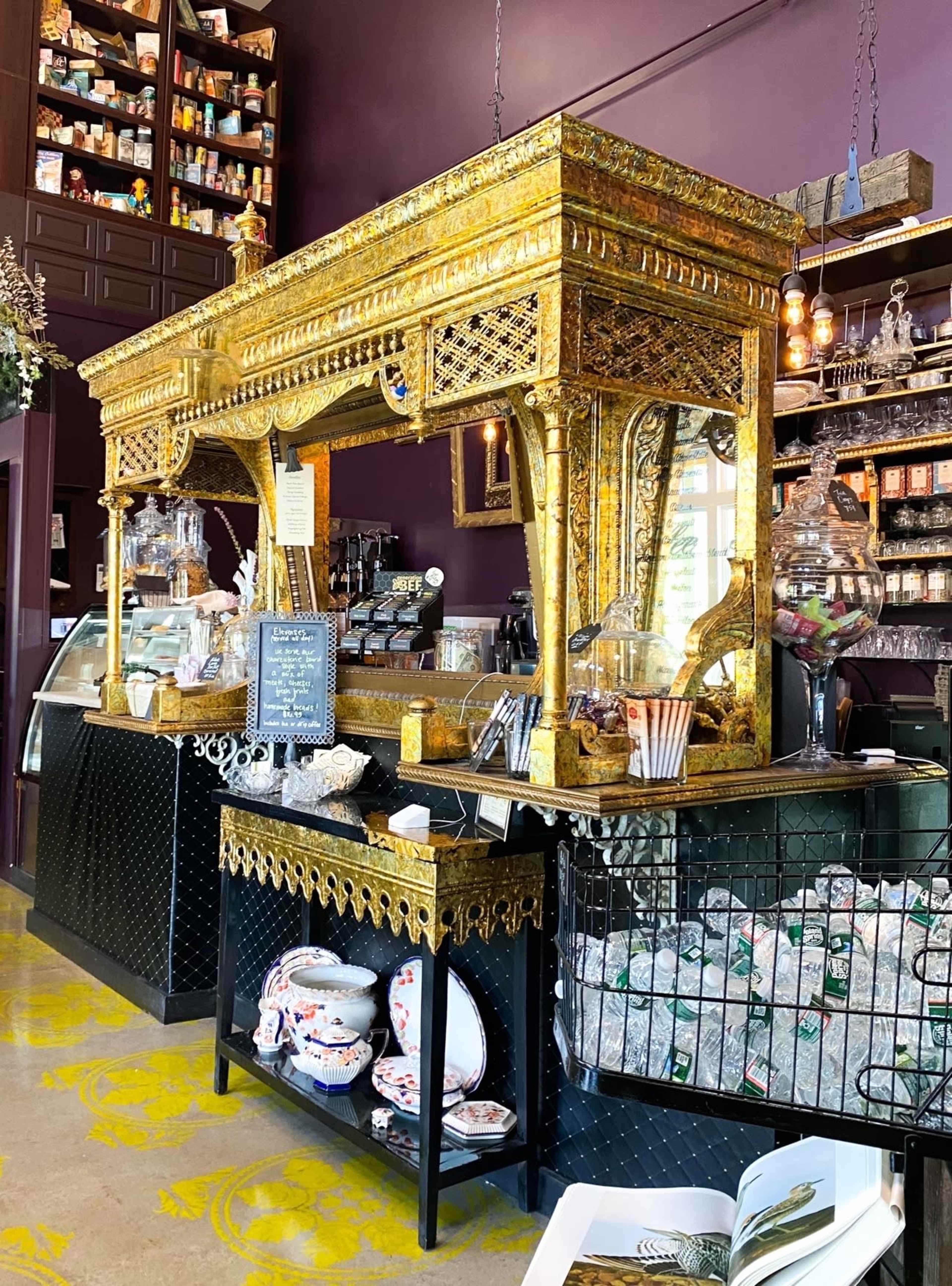 A richly decorated café counter featuring ornate golden accents, a glass display case, and shelves stocked with various jars and products in a dimly lit space.