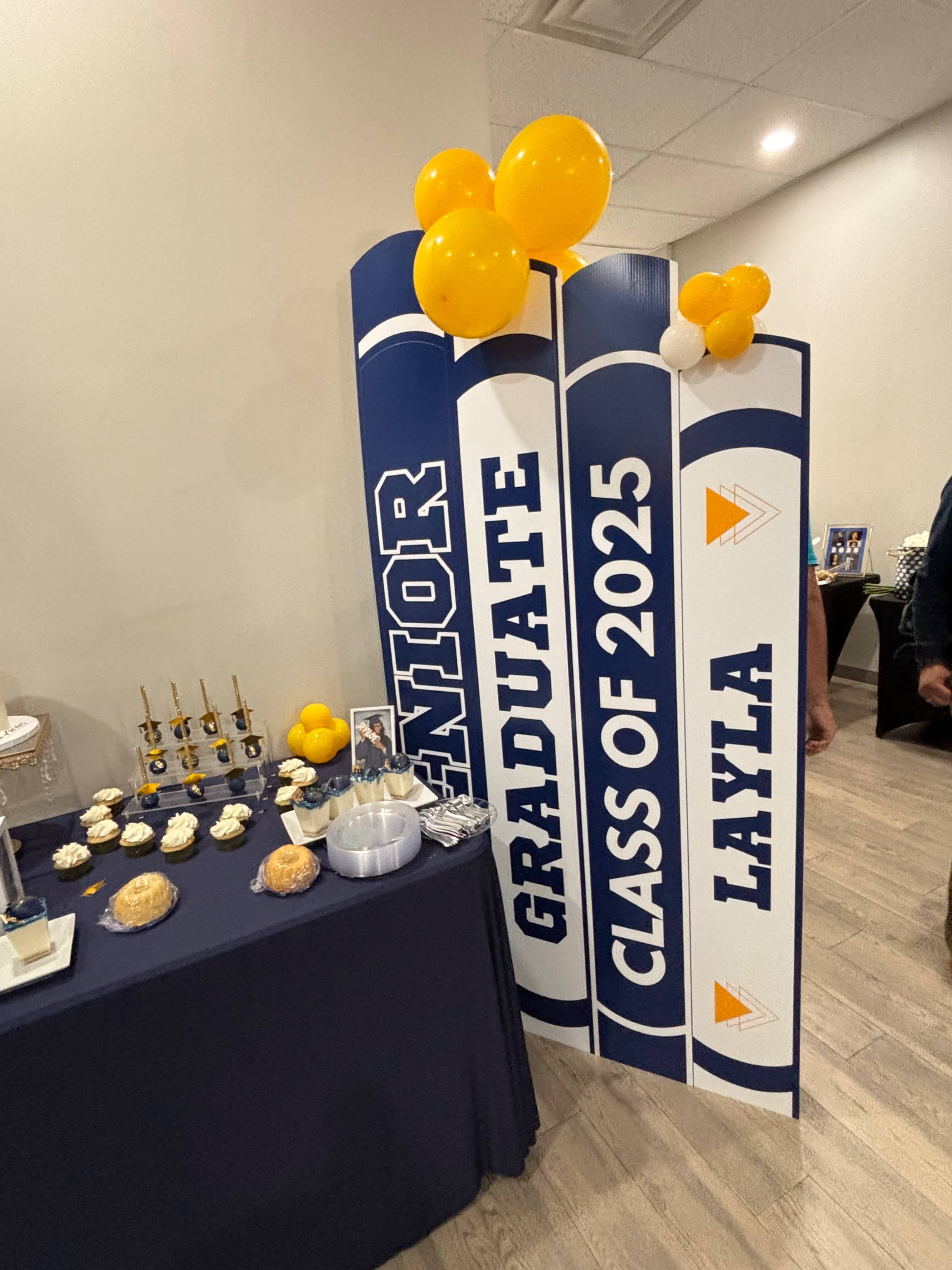 The image features a decorated table set for a graduation celebration, showcasing a large three-section display that reads "SENIOR," "GRADUATE," and "CLASS OF 2025," along with the name "LAYLA," and is adorned with yellow balloons.