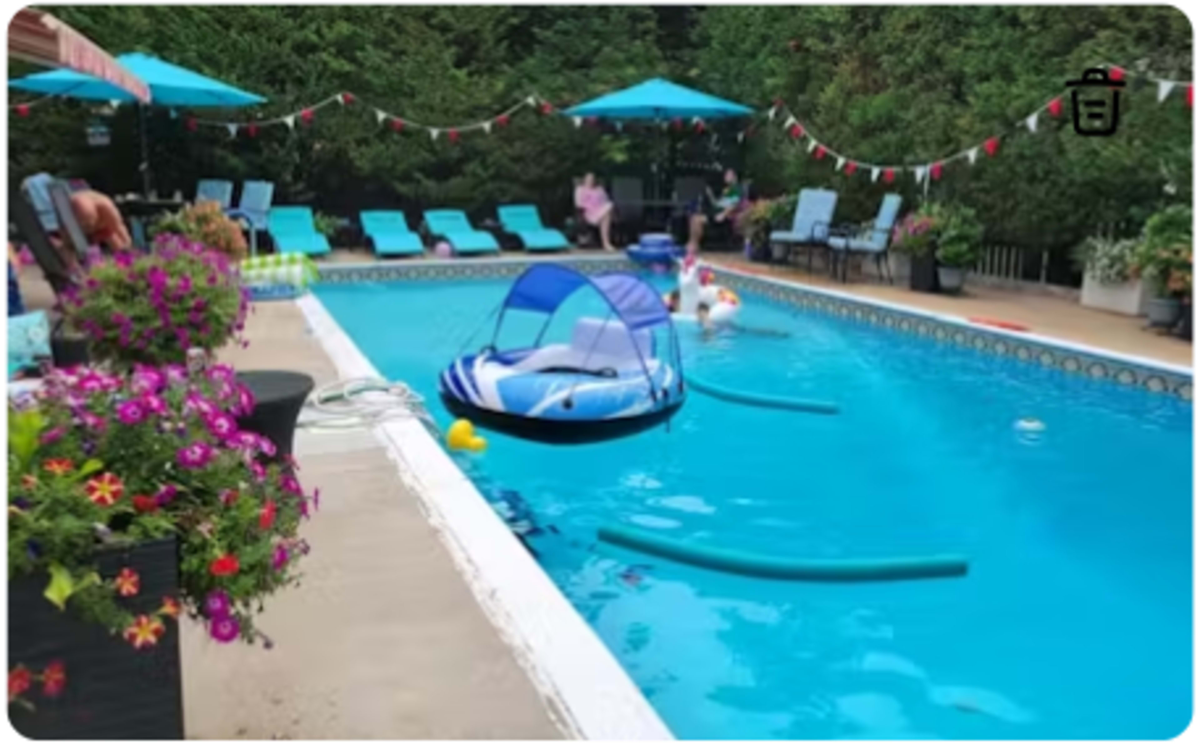 The image shows a swimming pool surrounded by lounge chairs, umbrellas, and colorful flowers, with pool floats and noodles scattered in the water.