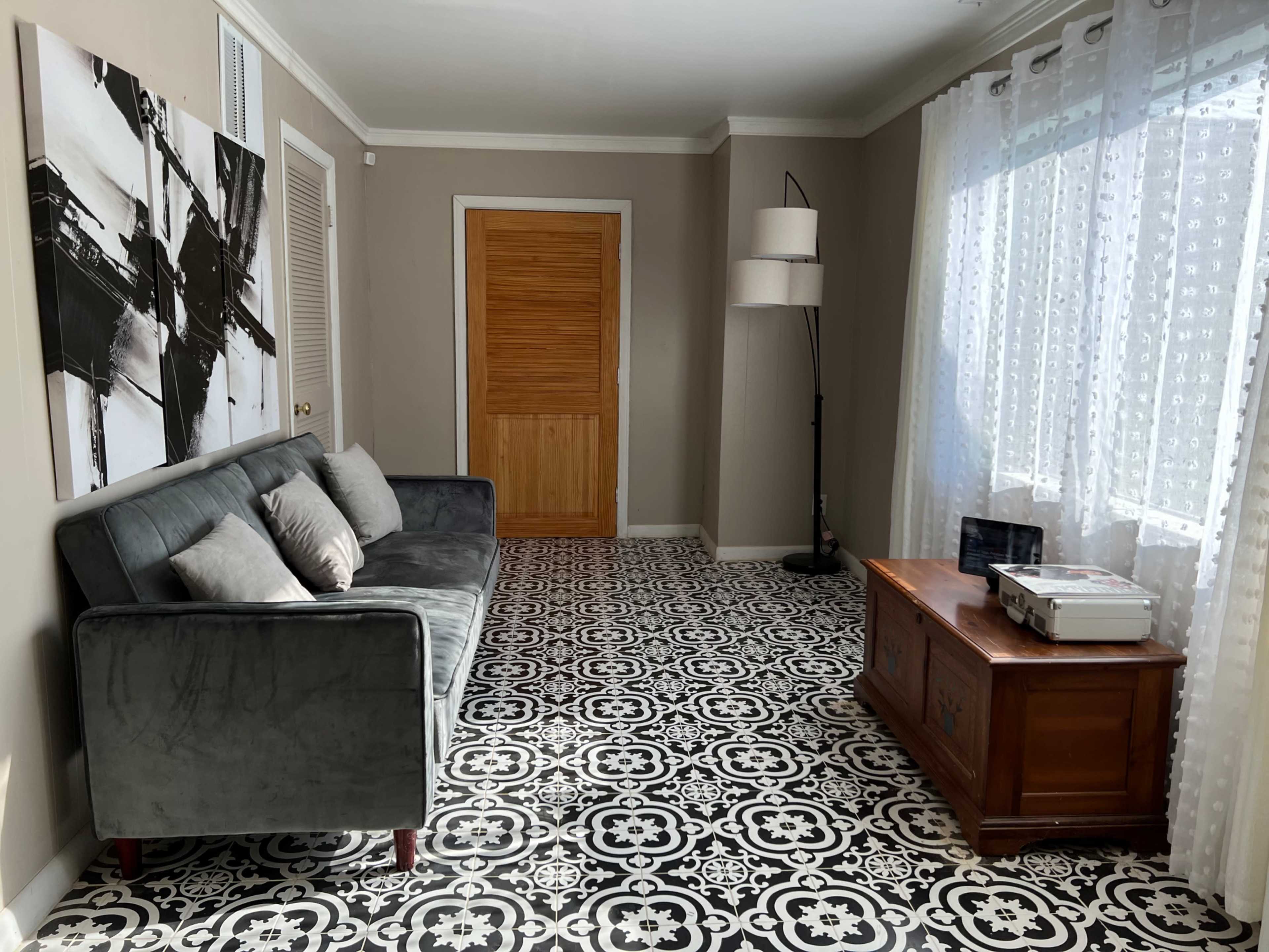A living room with a gray sofa, patterned floor tiles, a wooden cabinet, and a window with sheer curtains.