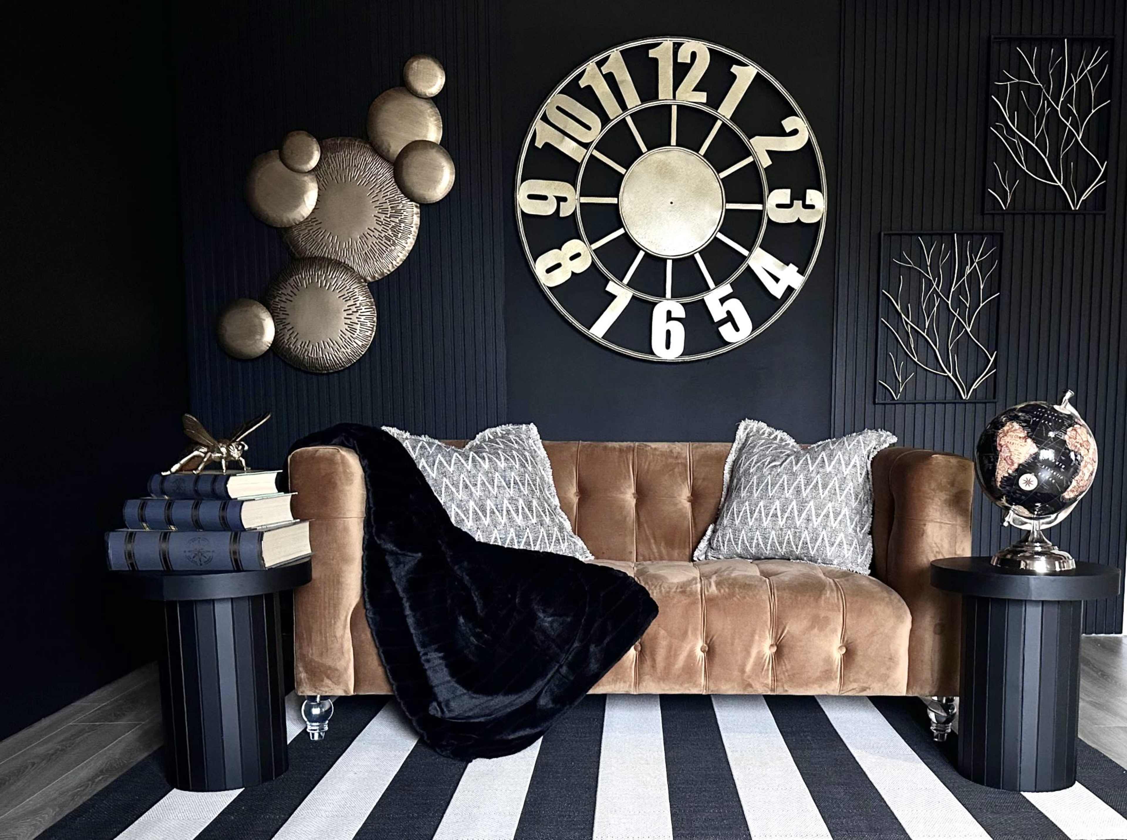 A brown couch with decorative pillows sits between two black side tables, featuring a large wall clock and metallic art pieces against a dark background.
