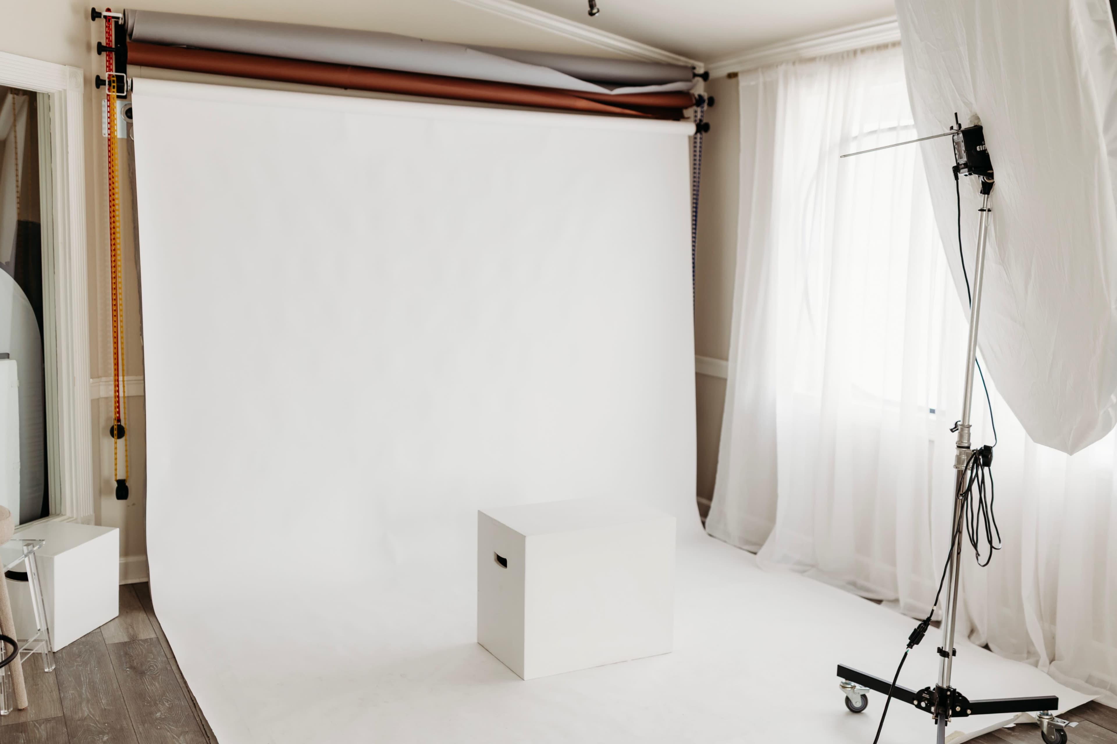 The image shows a photography studio with a white backdrop, a white cube on the floor, and sheer curtains letting in natural light.