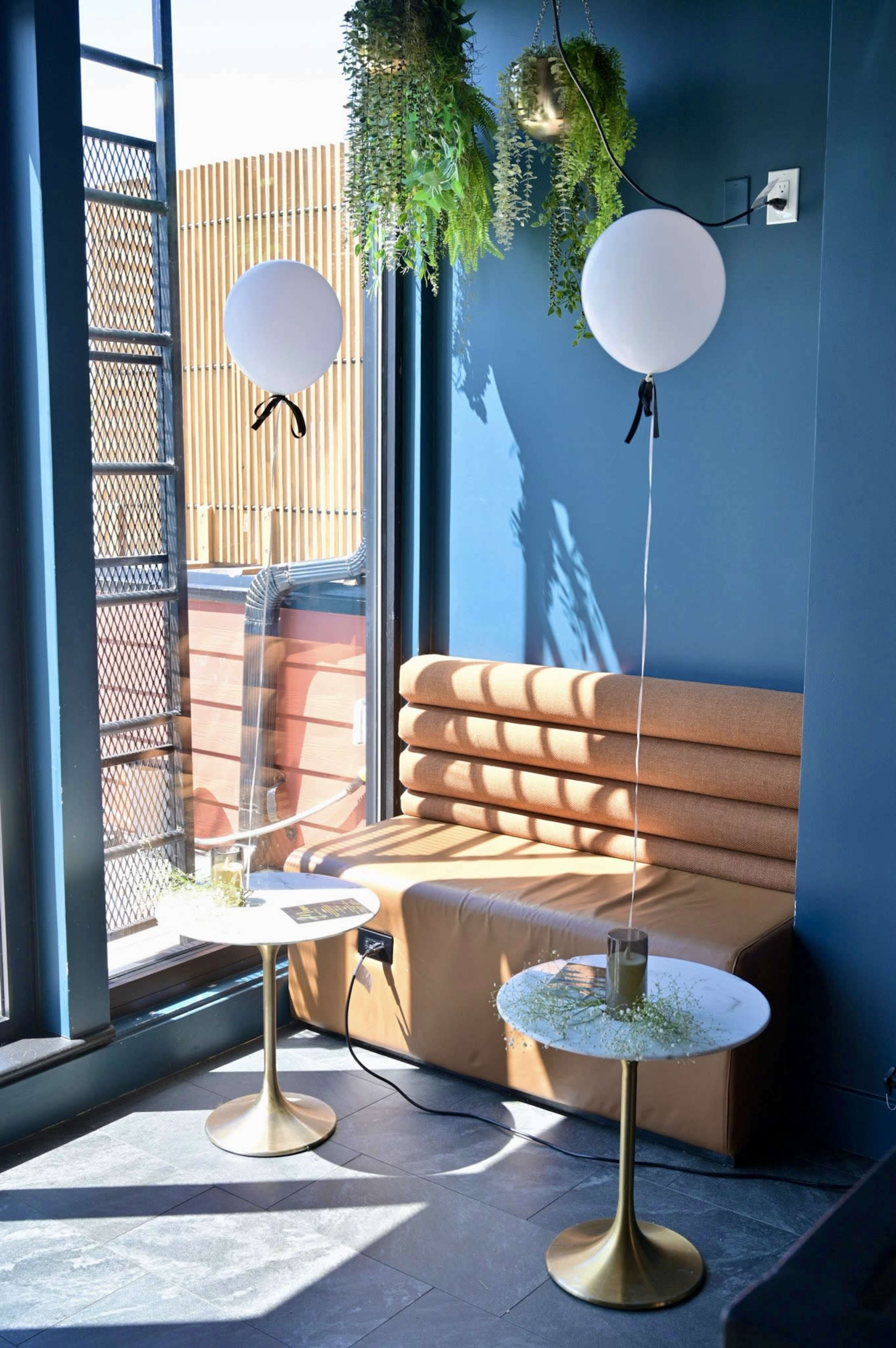 The image shows a cozy indoor seating area with a brown upholstered bench, two small round tables with gold bases, and two white balloons tied with black ribbons.