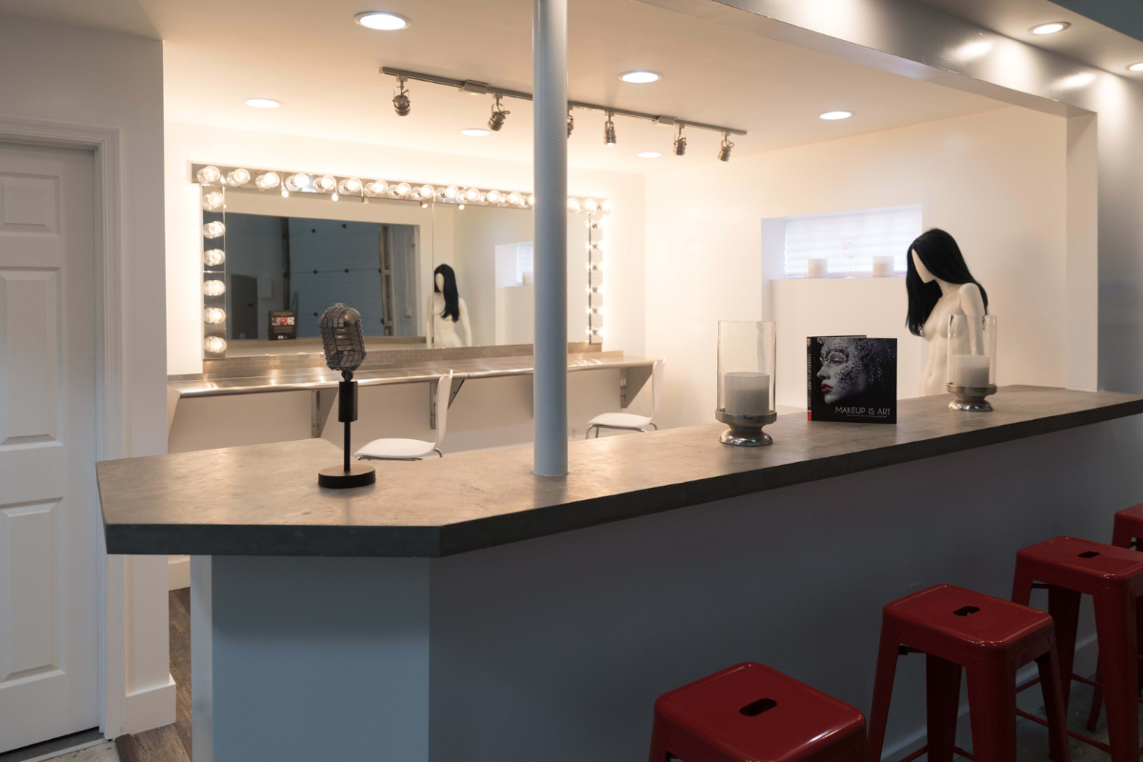 The image shows a modern bar area with a concrete countertop, stylish red stools, and a large mirror illuminated by bright lights.