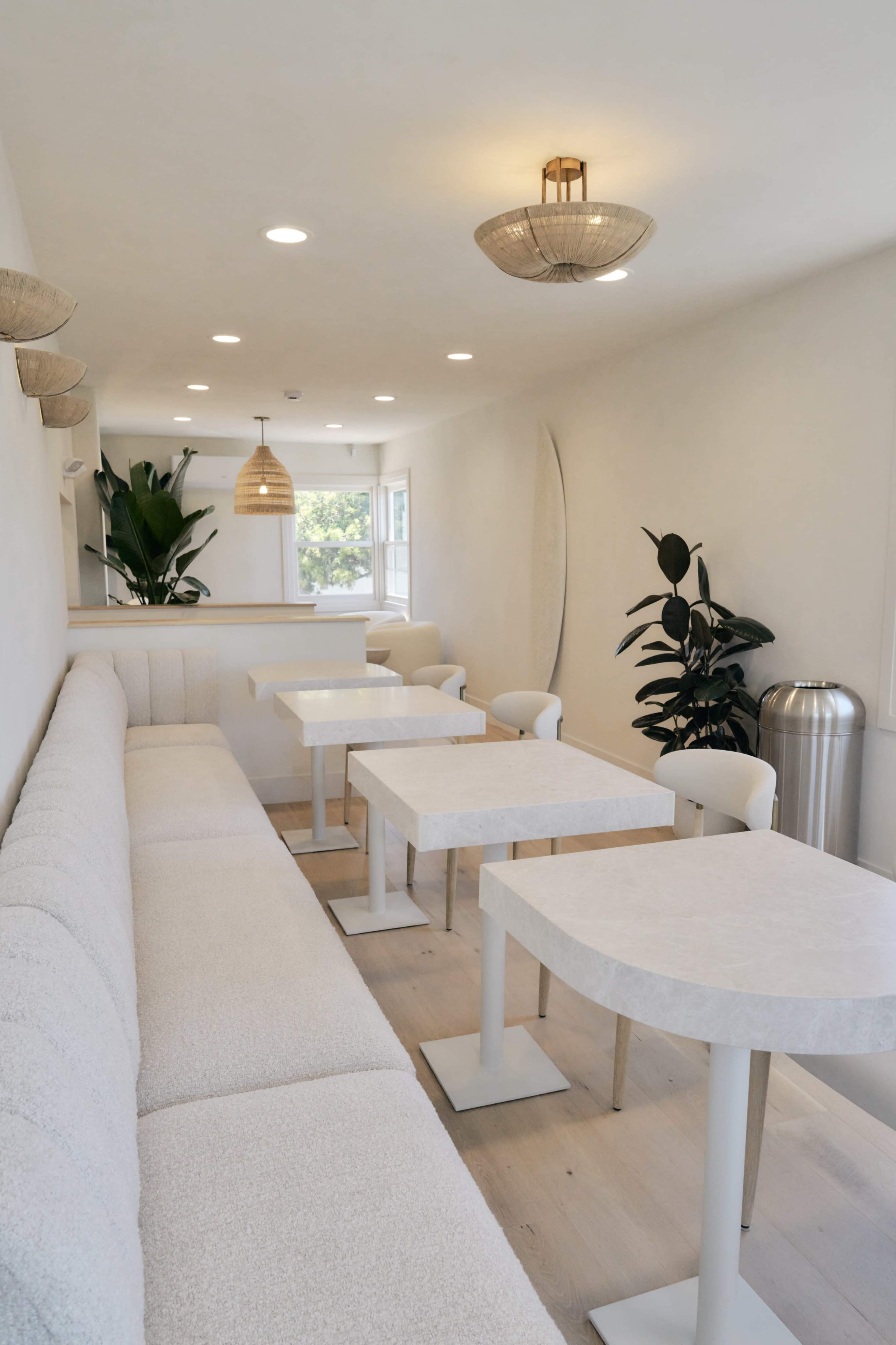 The image shows a bright, minimalist interior of a café or restaurant with white furniture and several small tables arranged along the walls.