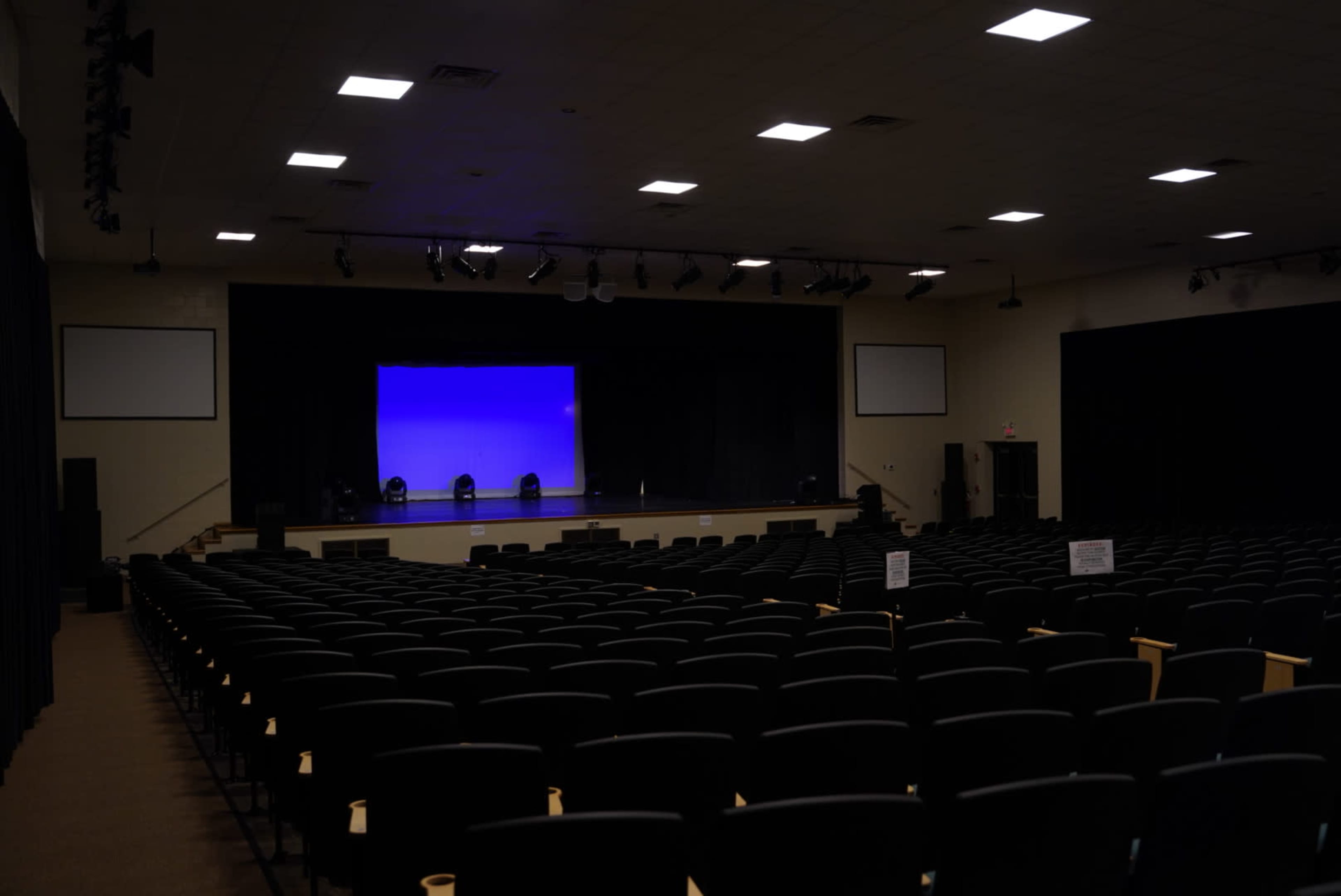 An empty auditorium with rows of black chairs facing a stage with a blue backdrop.