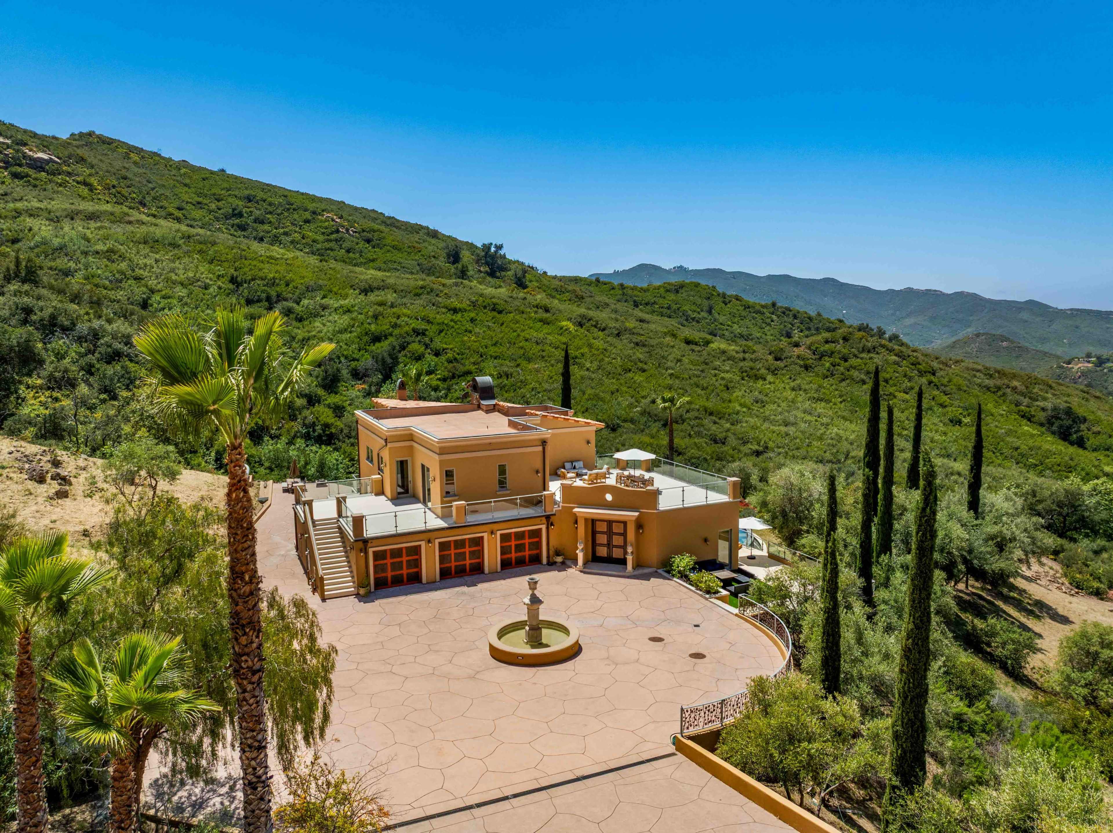 The image shows a large, two-story house surrounded by green hills, with a circular driveway and a fountain at the center.