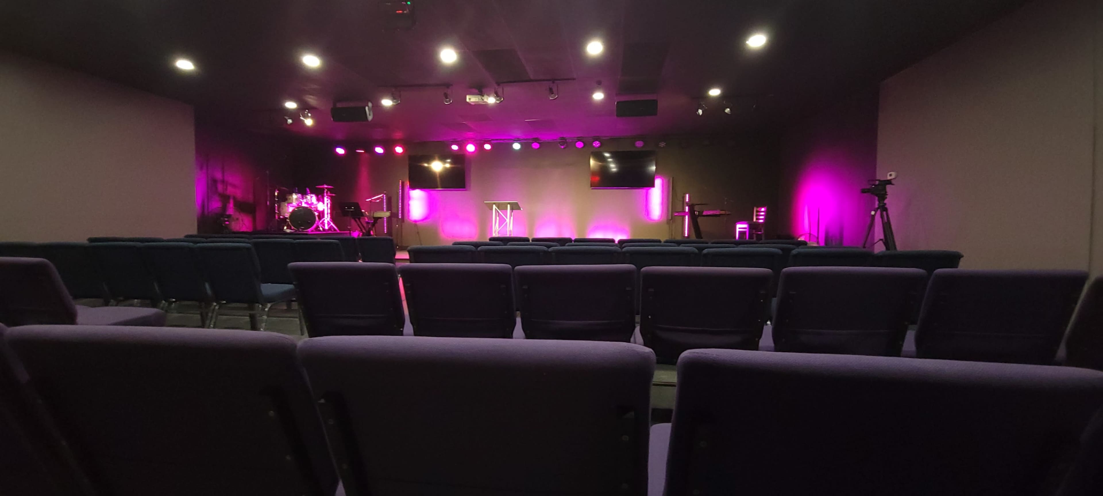 The image shows an empty auditorium with purple lighting, featuring a stage with a podium, musical instruments, and cameras set up for recording.