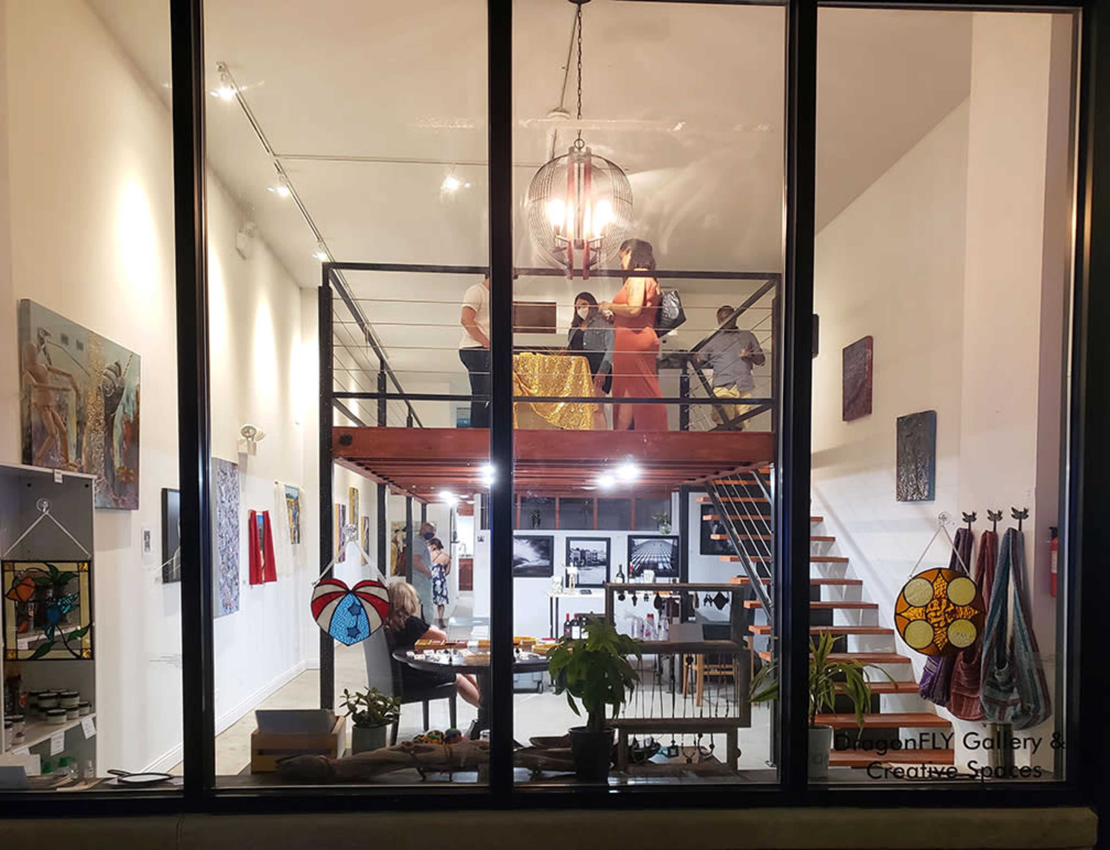 The image shows the interior of a gallery with people interacting and artwork displayed on the walls, viewed through large glass windows.