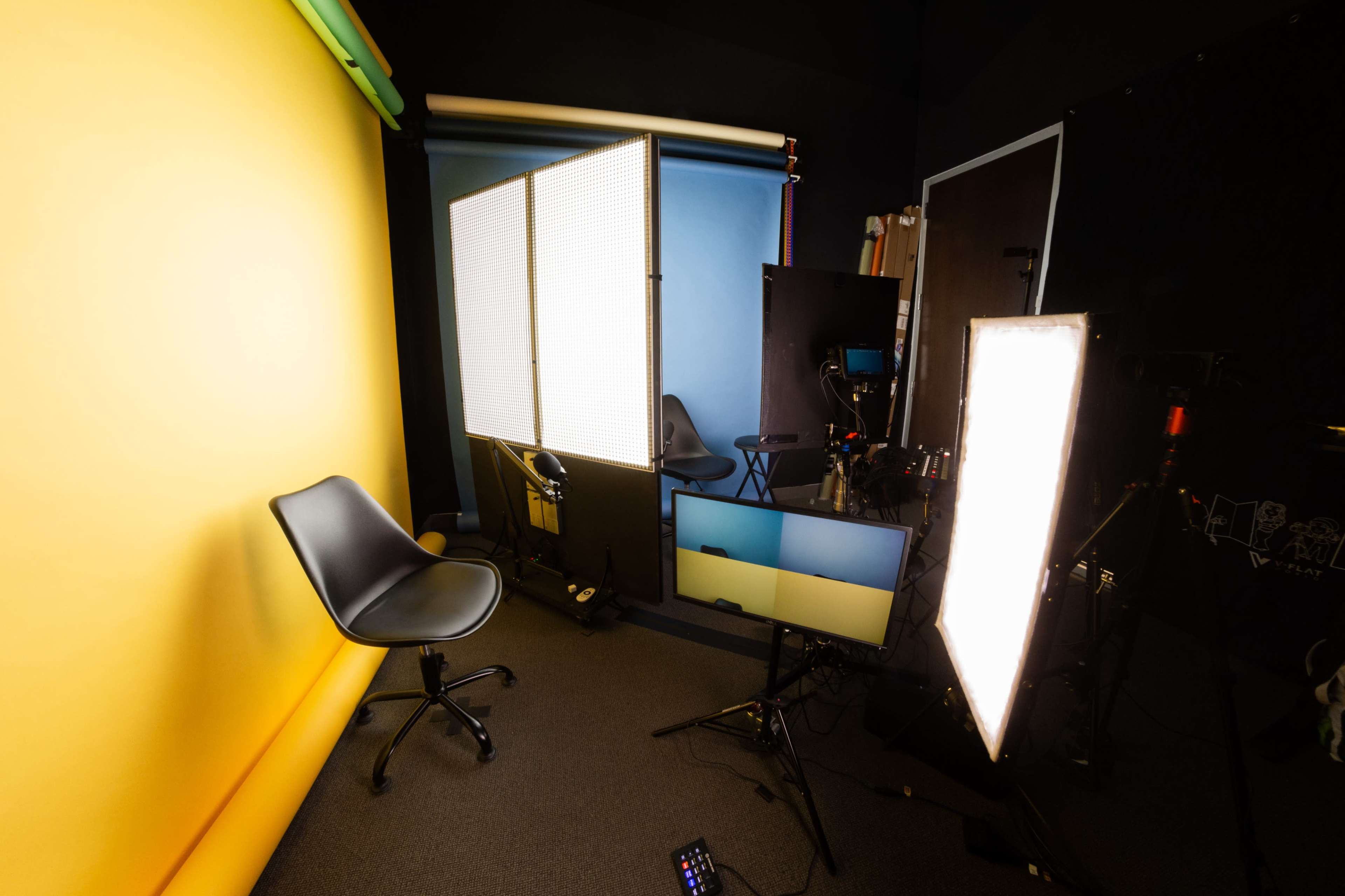A studio setup features a black chair in front of a yellow backdrop, with a blue backdrop visible and various lighting and equipment arranged nearby.