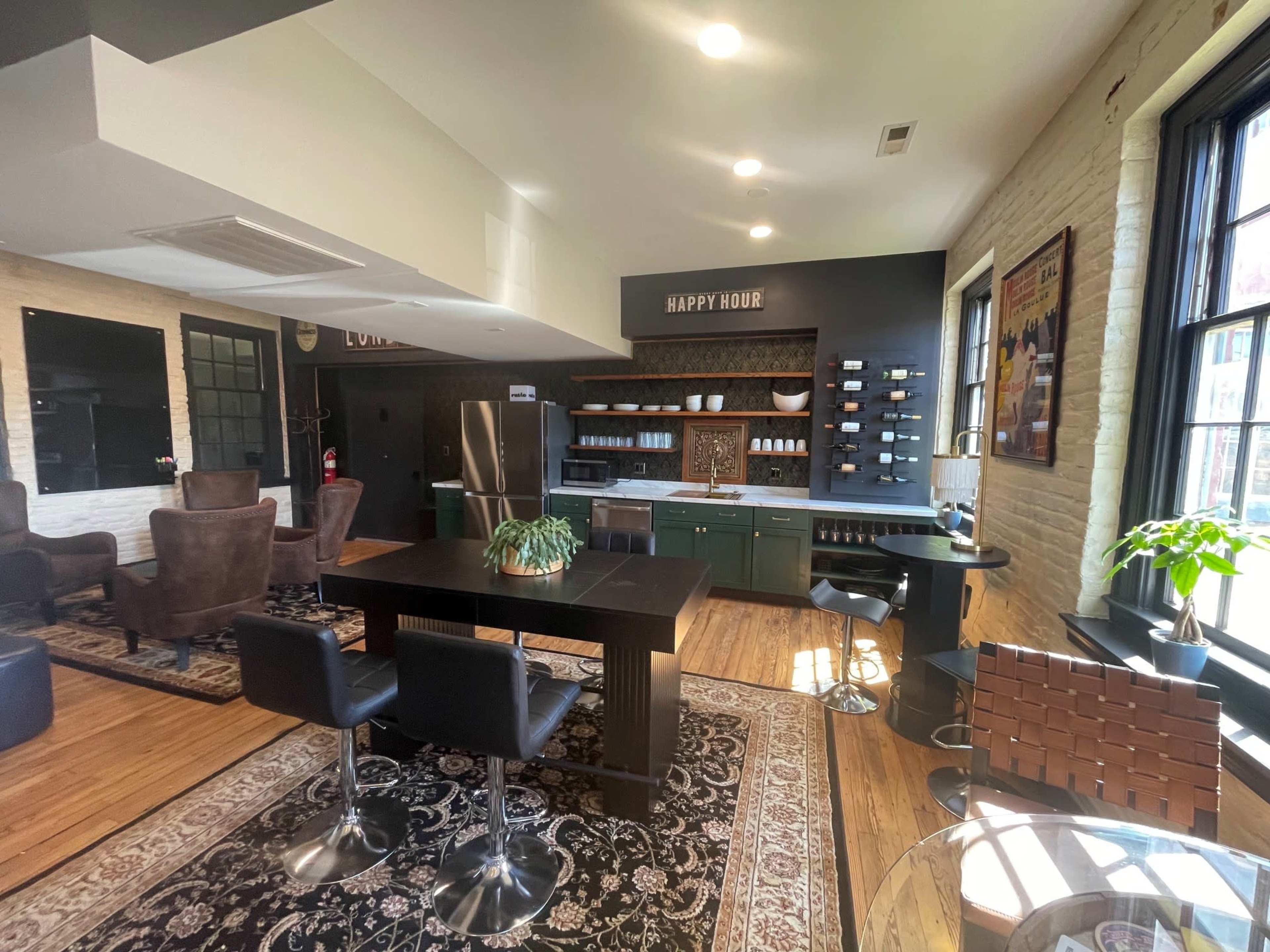 The image shows a stylish open kitchen and lounge area featuring modern furniture, a large table with bar stools, a refrigerator, and shelves with neatly arranged dishes.