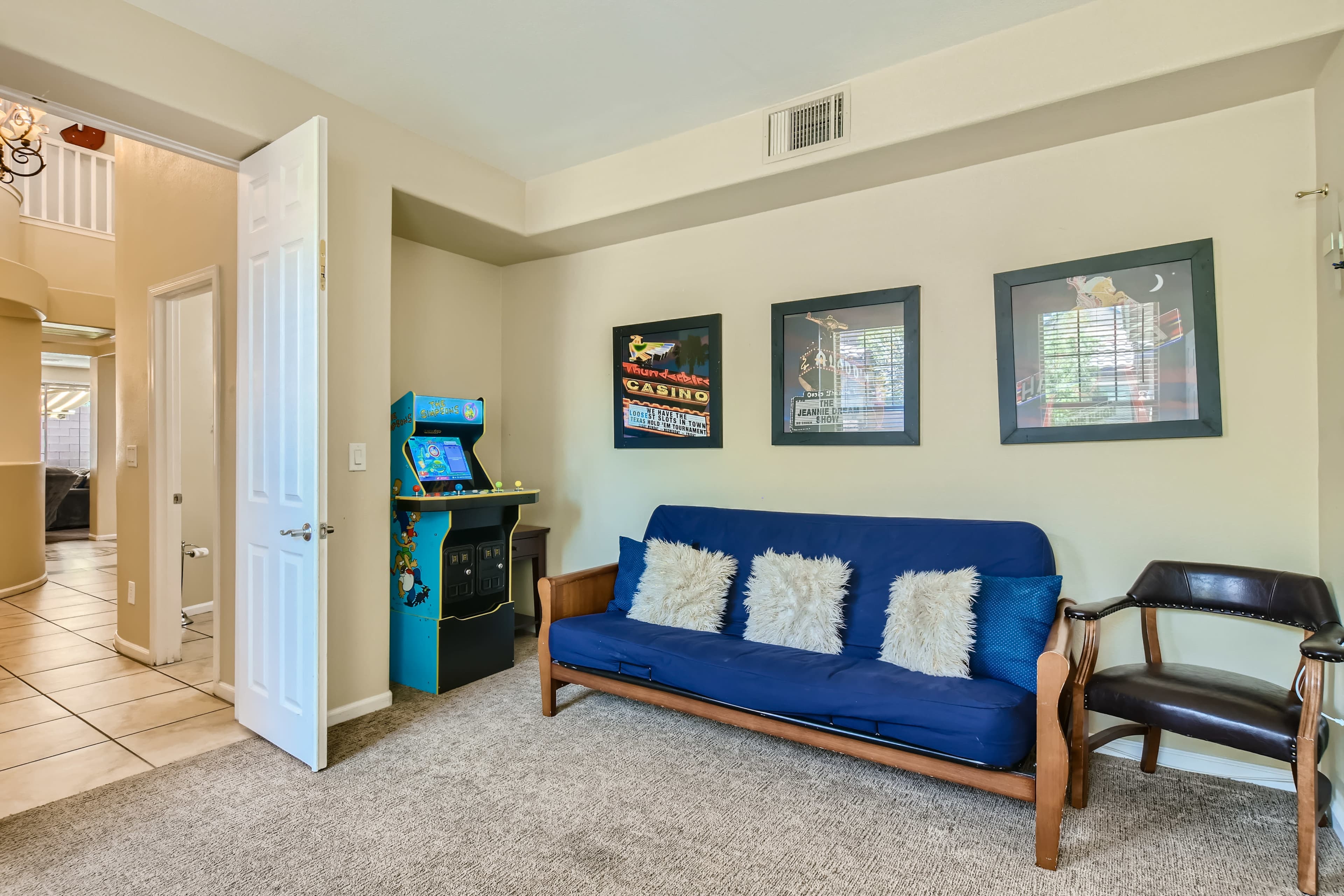 A cozy living room with a blue futon, a black chair, framed art on the walls, and a vintage arcade game machine in the corner.