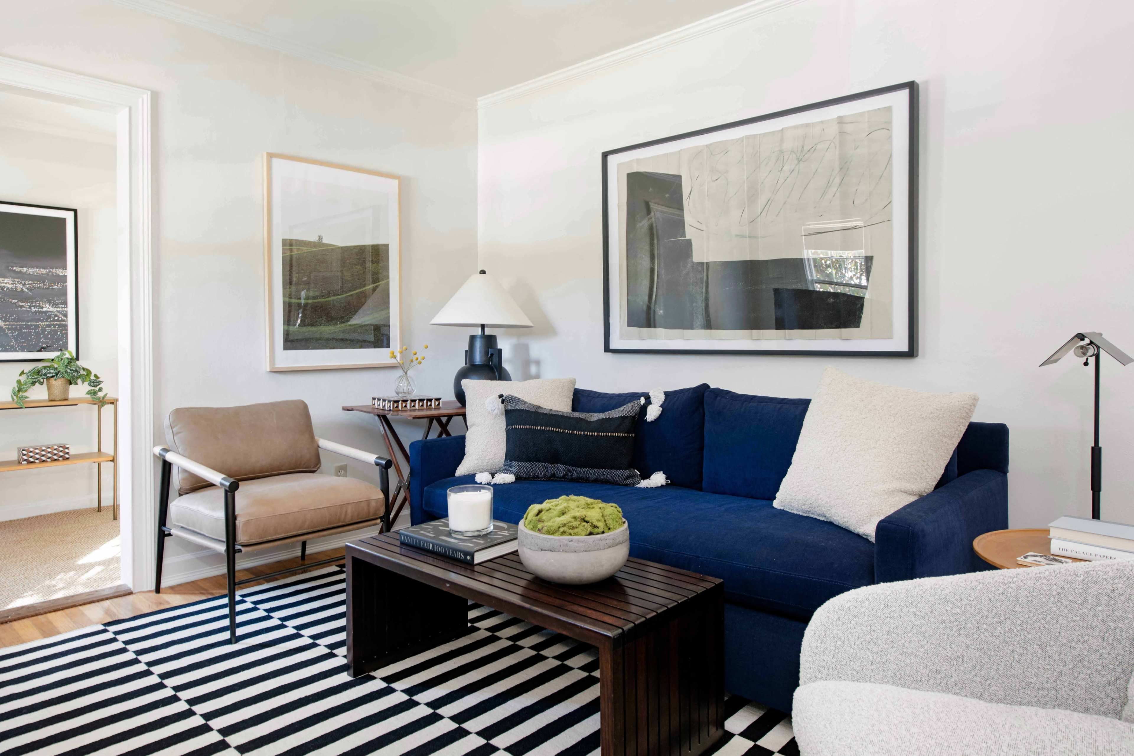 A living room with a blue sofa, a wooden coffee table, an armchair, a lamp, framed artwork on the walls, and a striped rug.