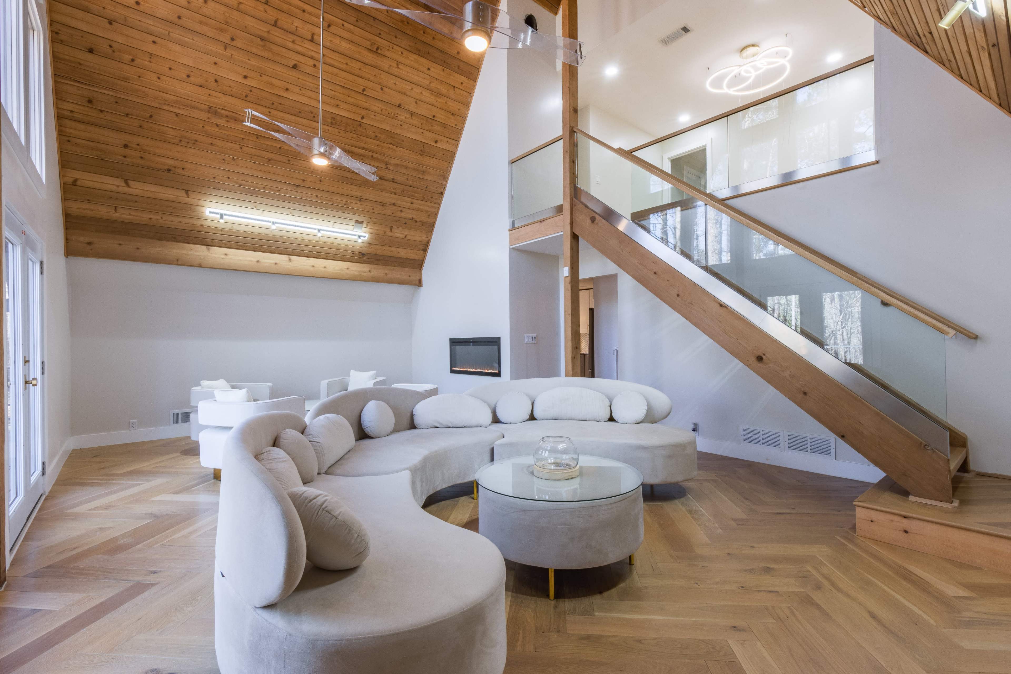 A modern living room with a curved sofa, a glass coffee table, and a staircase leading to an upper level, all accented by wood paneling and large windows.