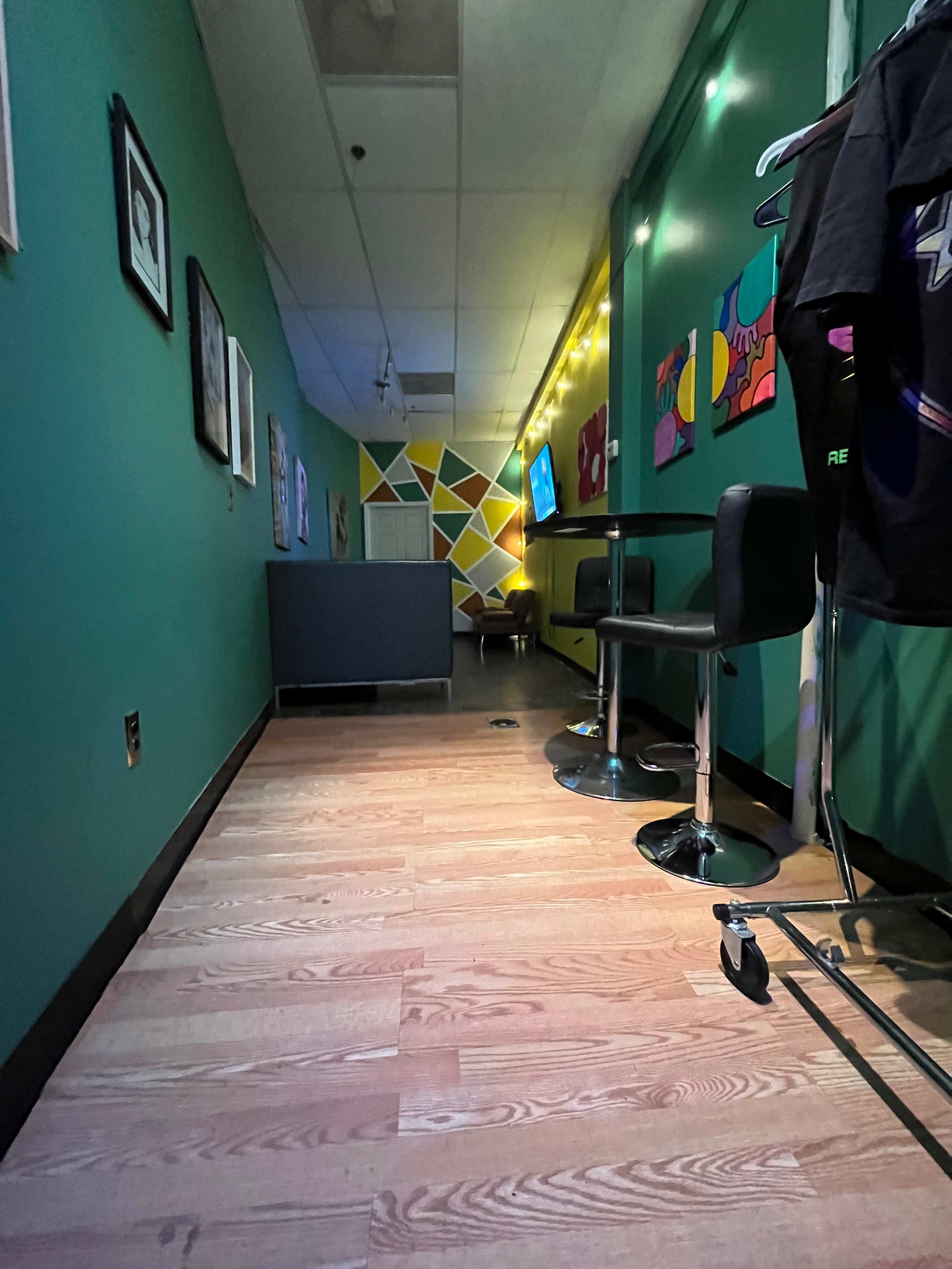 The image shows a hallway with green walls, wooden flooring, bar stools, and colorful artwork on the walls.