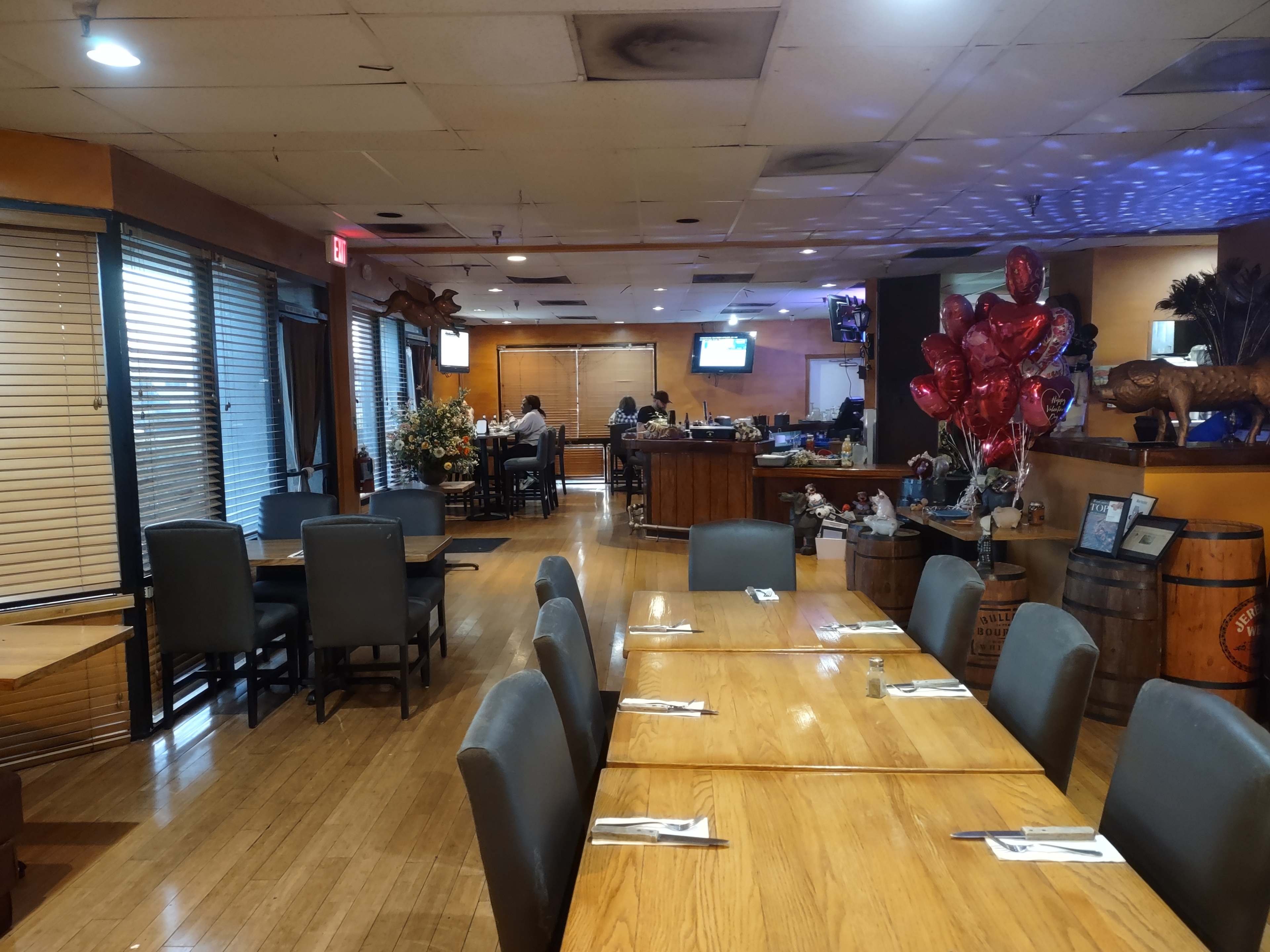 A restaurant interior with wooden tables and chairs, decorations, and a bar area in the background.