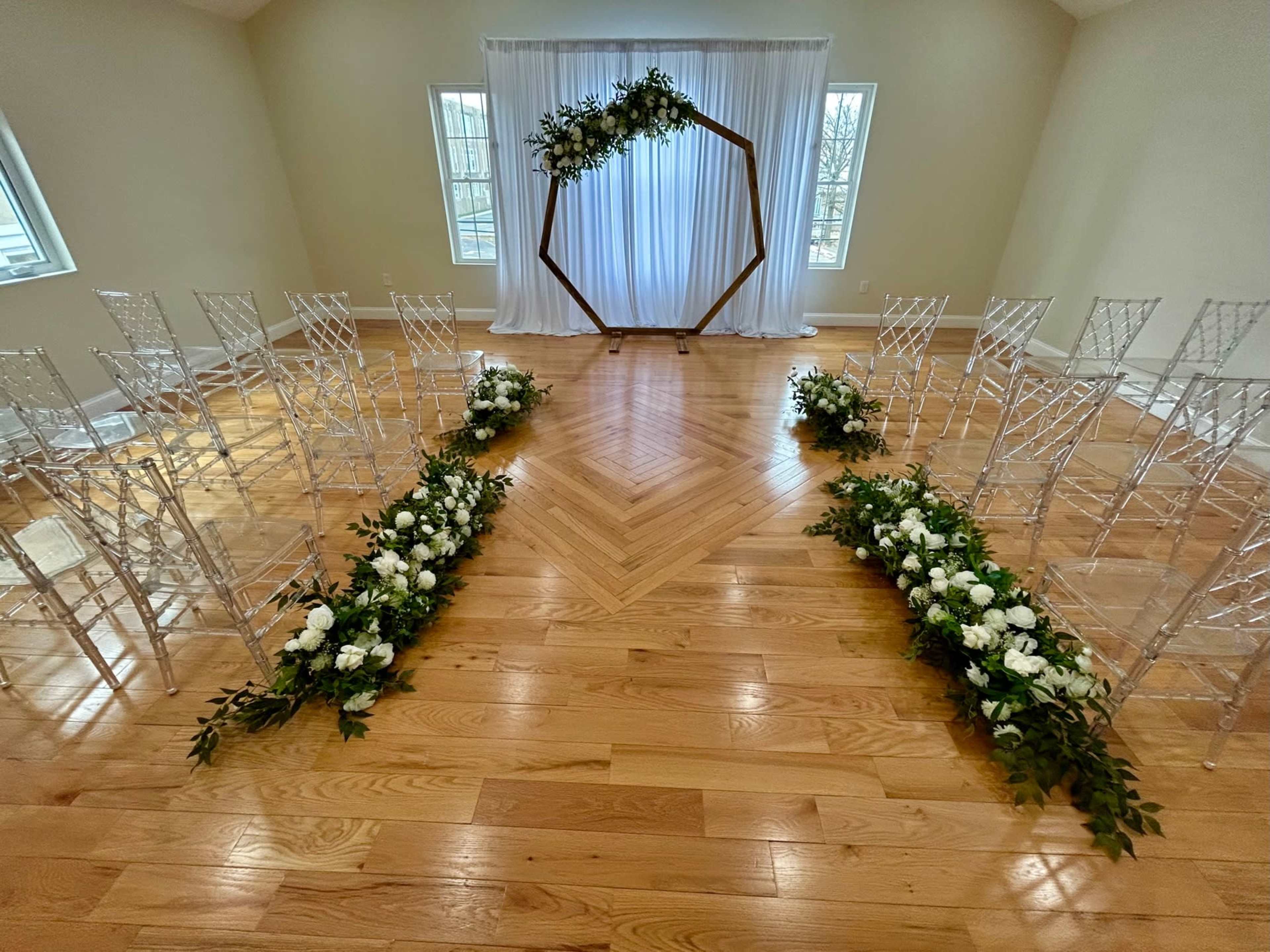 The image shows a wedding ceremony setup with transparent chairs arranged in two rows facing an ornate wooden arch decorated with greenery and white flowers, in front of a backdrop of flowing white fabric.