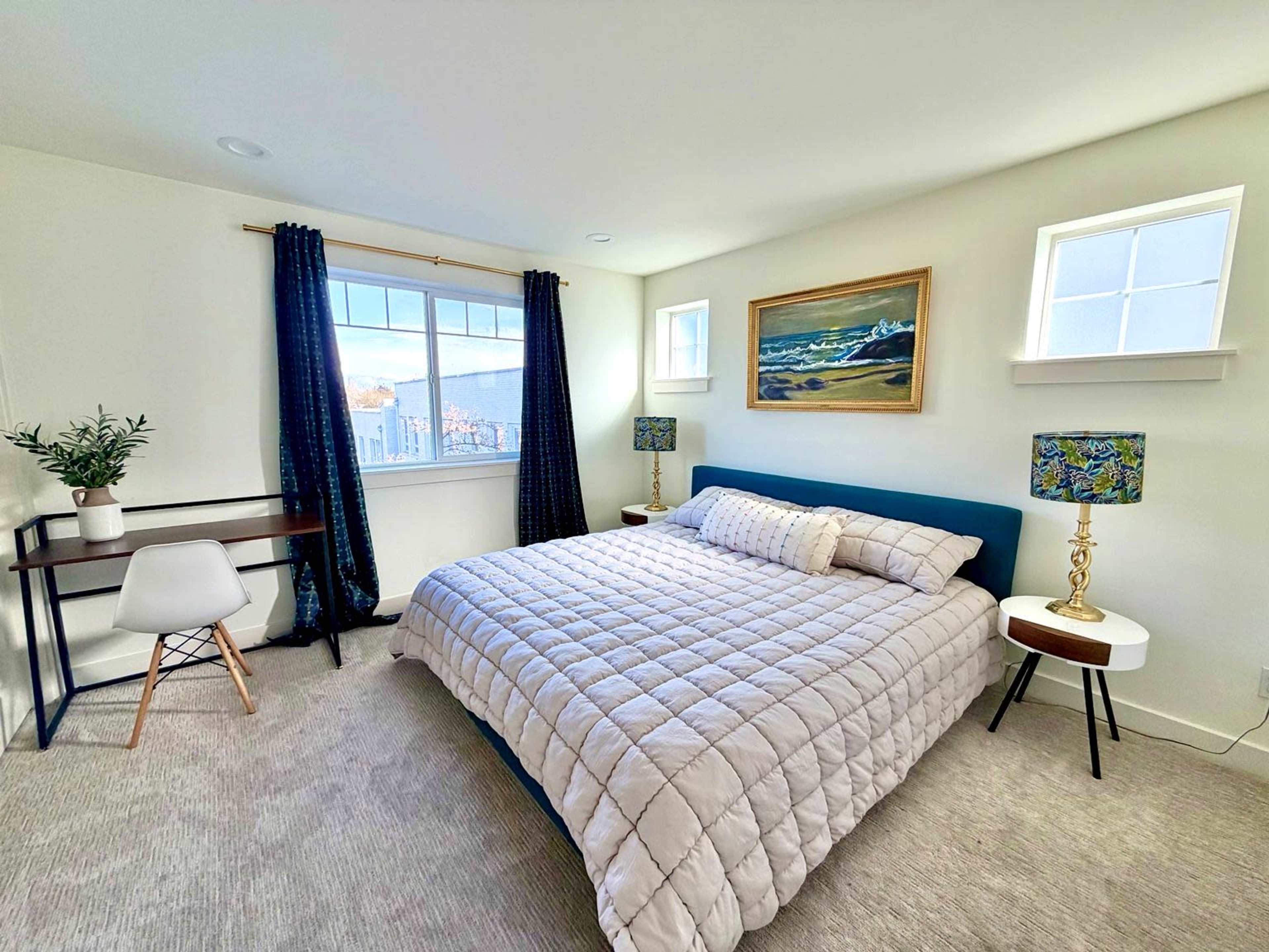 The image shows a cozy bedroom with a large bed, two side tables with lamps, a desk, and a window overlooking an outdoor view.
