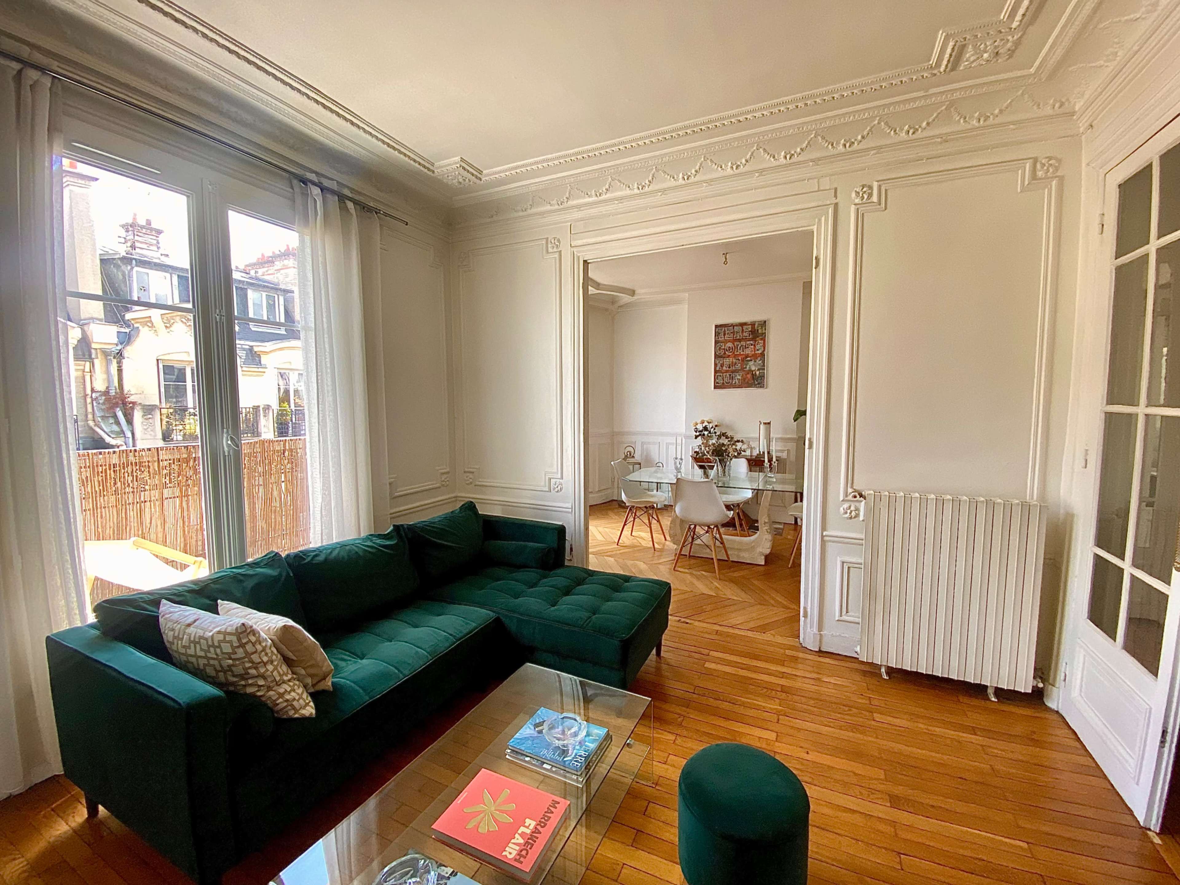 The image shows a well-lit living room featuring a green sectional sofa, a coffee table with magazines, and a view of a dining area through an open doorway.