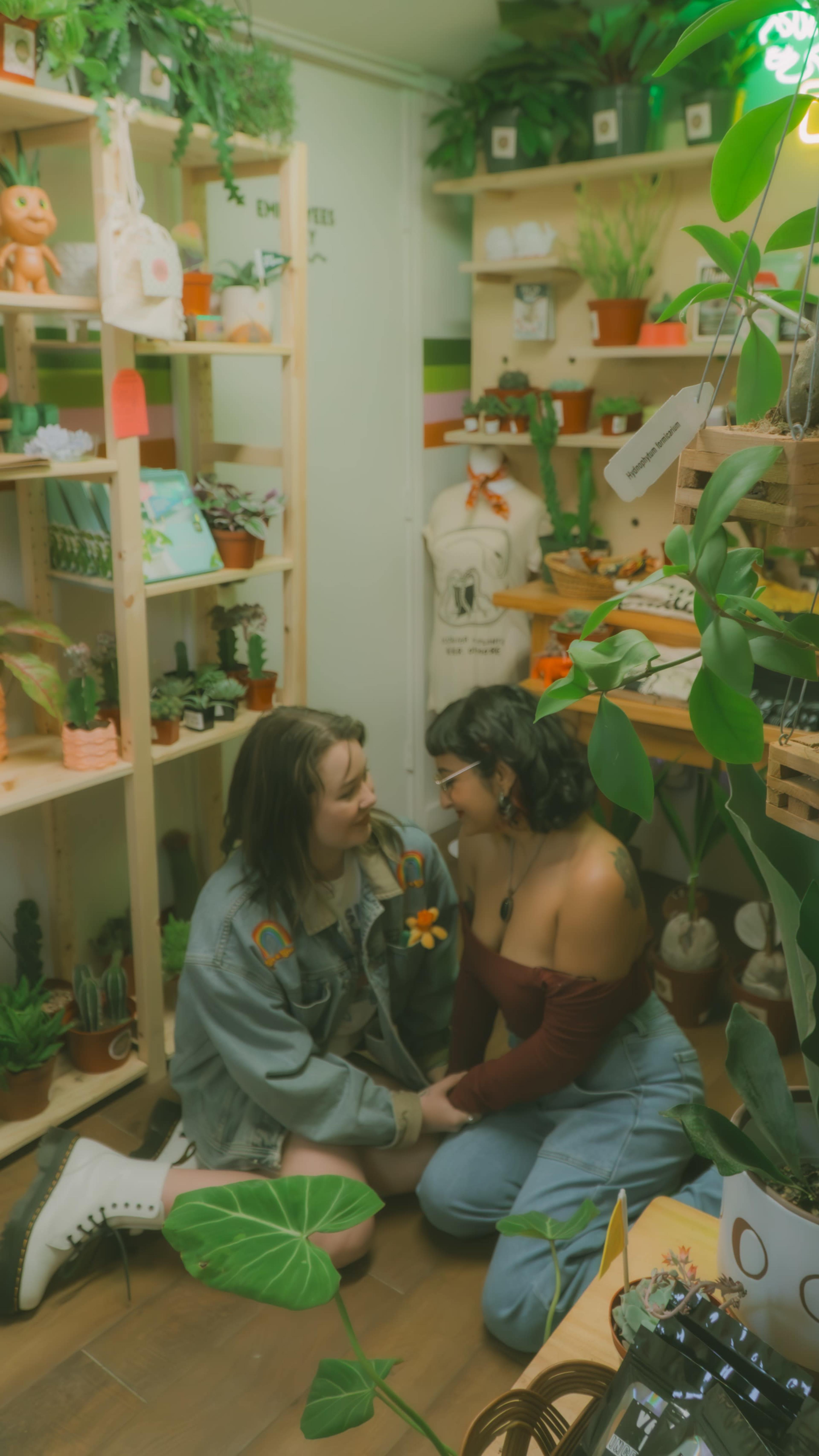 Two women sit on the floor of a plant-filled shop, surrounded by shelves of greenery and decorative items.