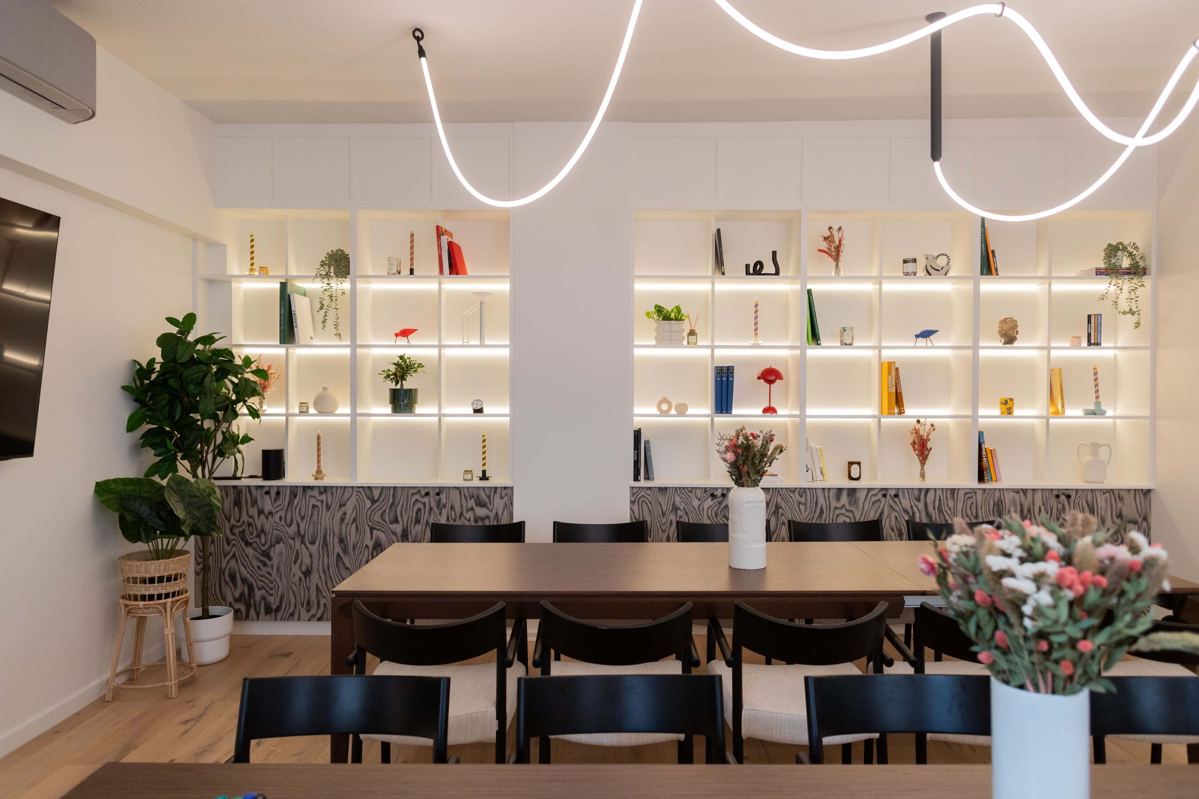 A modern meeting room features a long wooden table with black chairs and a wall of illuminated shelves displaying decorative items and plants.