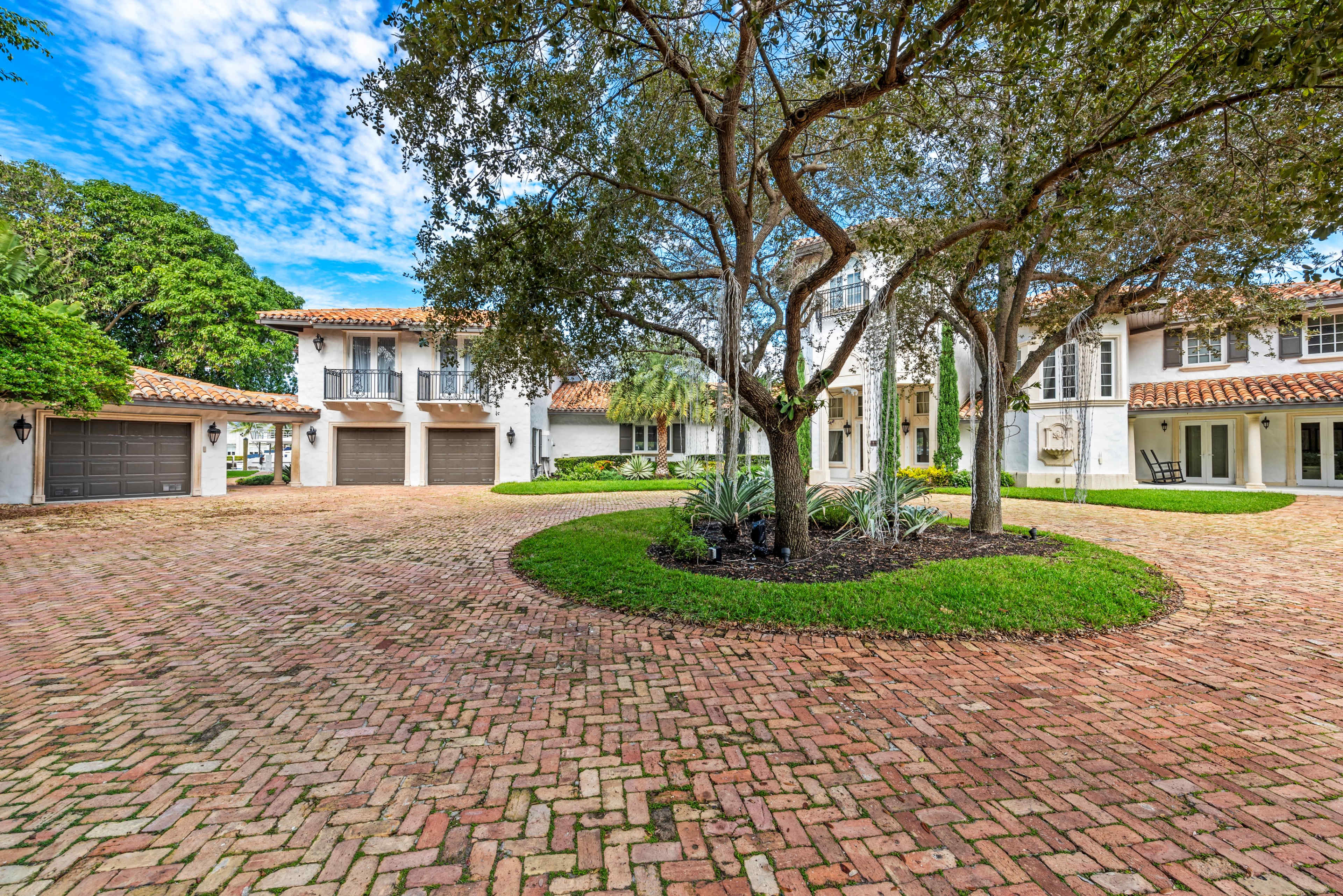 The image shows a residential property with a circular driveway, landscaped greenery, and garage doors on either side of the house.