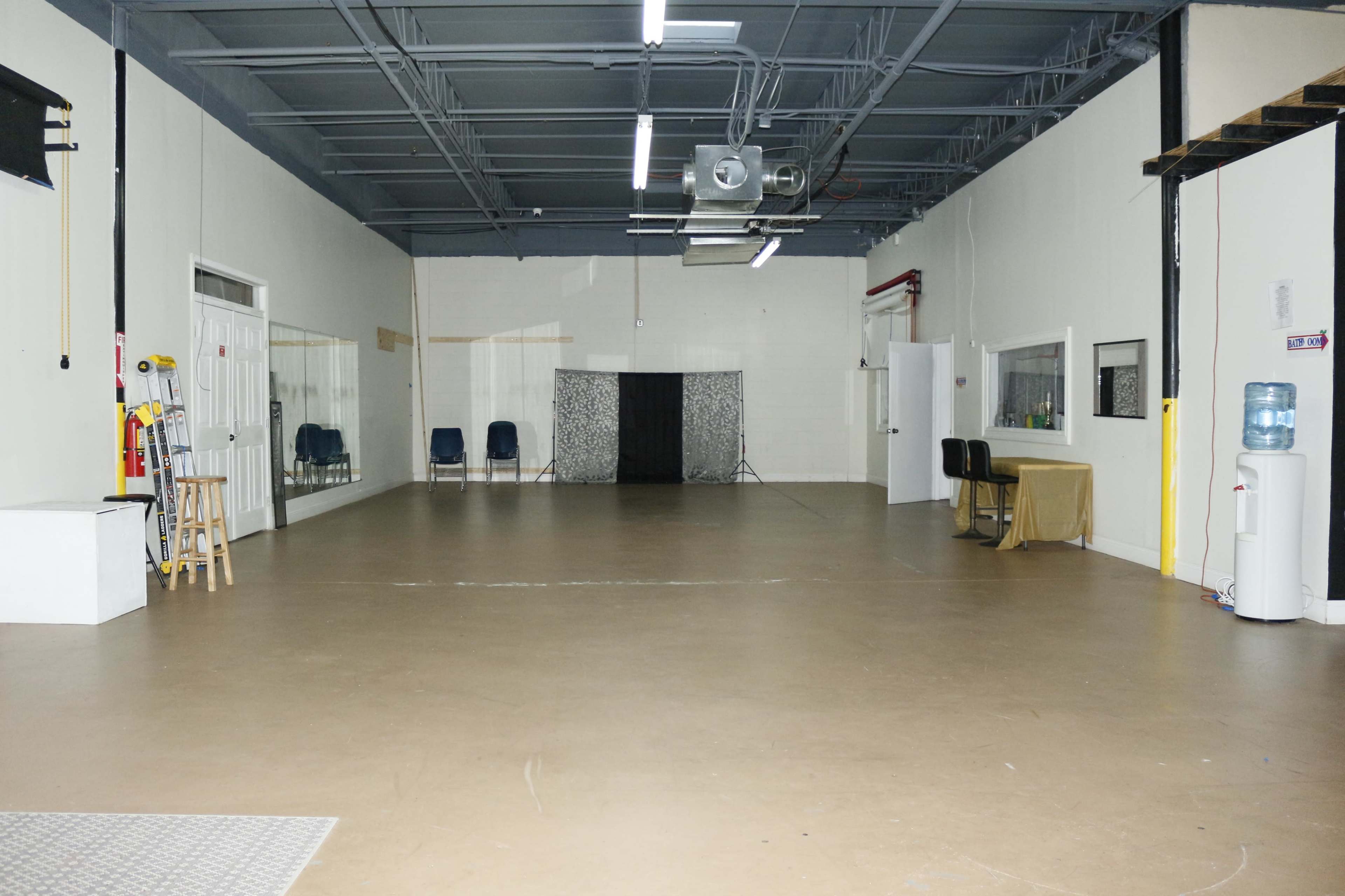A spacious, empty indoor area with a light-colored floor, white walls, and minimal furniture, including two black chairs and a table.