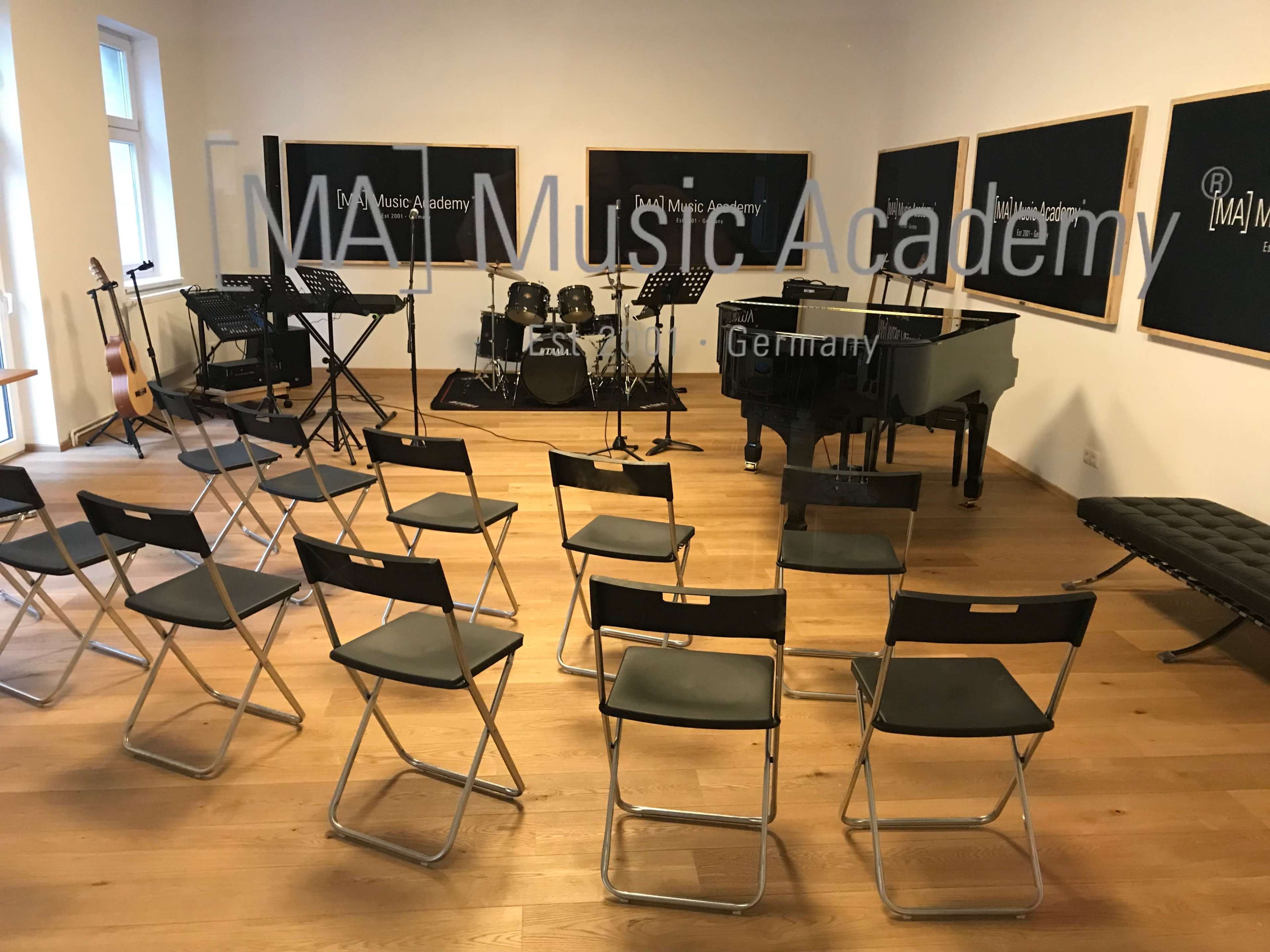 A music rehearsal room at the MA Music Academy in Germany features an empty seating area facing a stage equipped with a grand piano, drums, and electronic instruments.