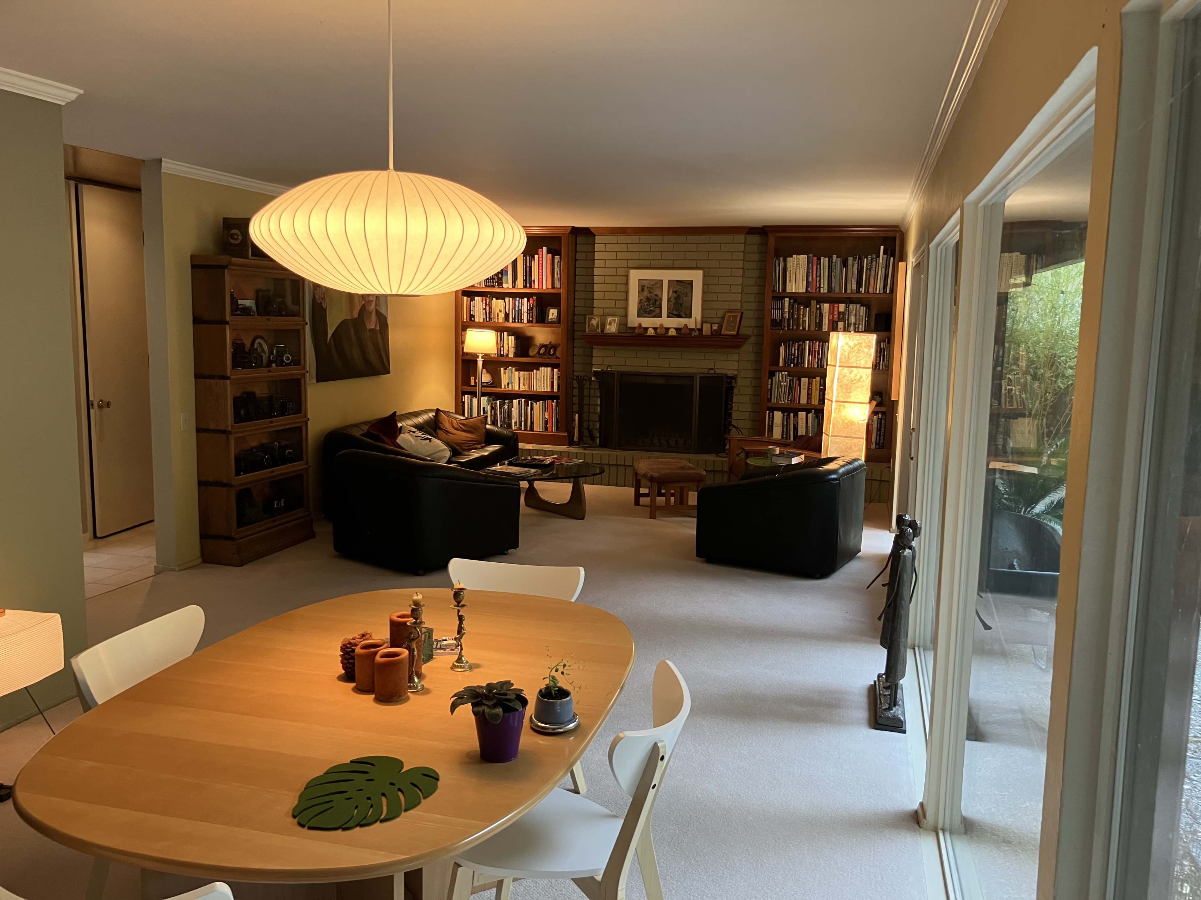 A living space featuring a dining table with chairs, a bookshelf-filled wall, a black sofa, and large windows allowing light to enter.