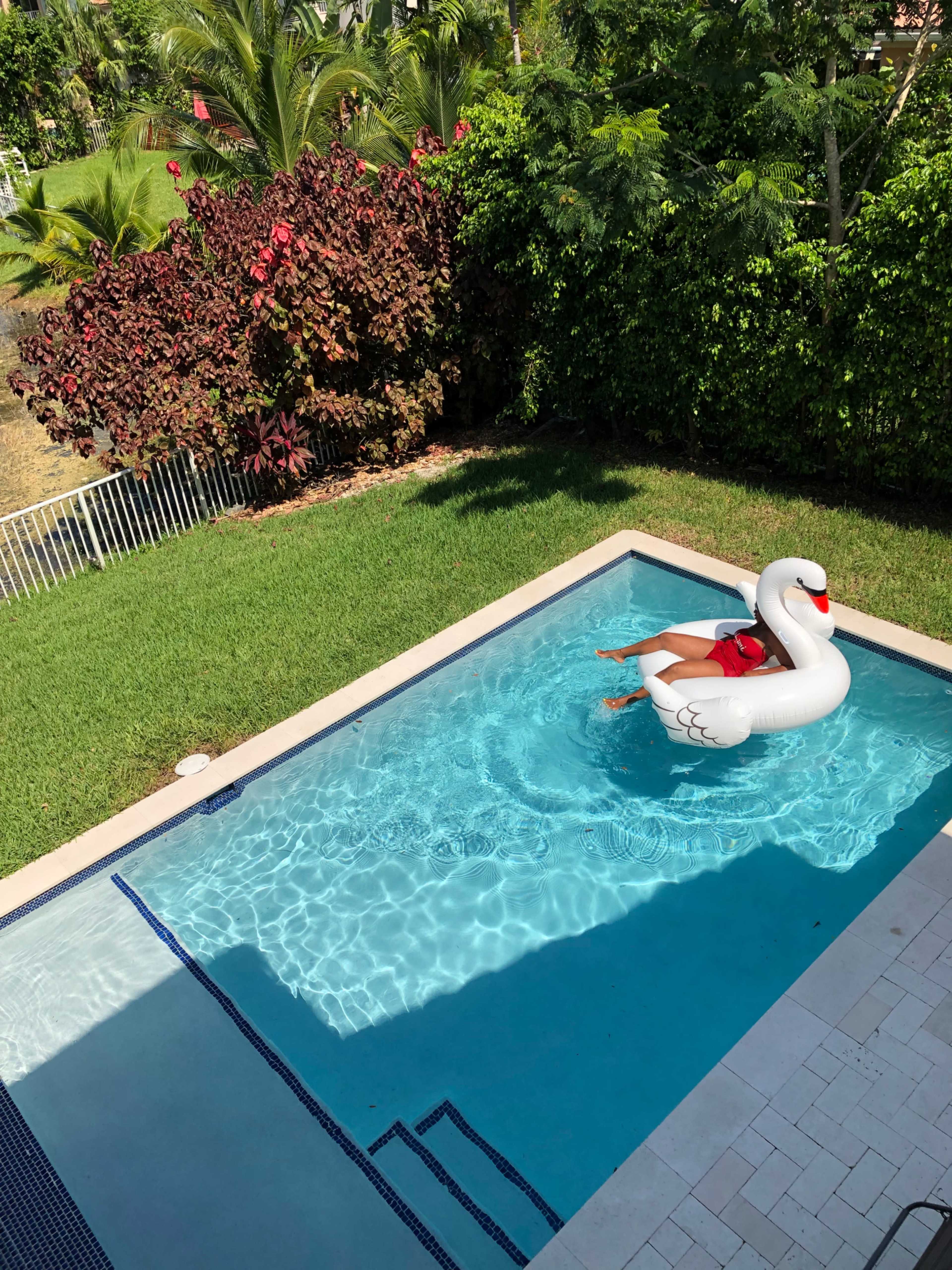 A person wearing a red swimsuit relaxes on a large white swan float in a swimming pool surrounded by greenery.