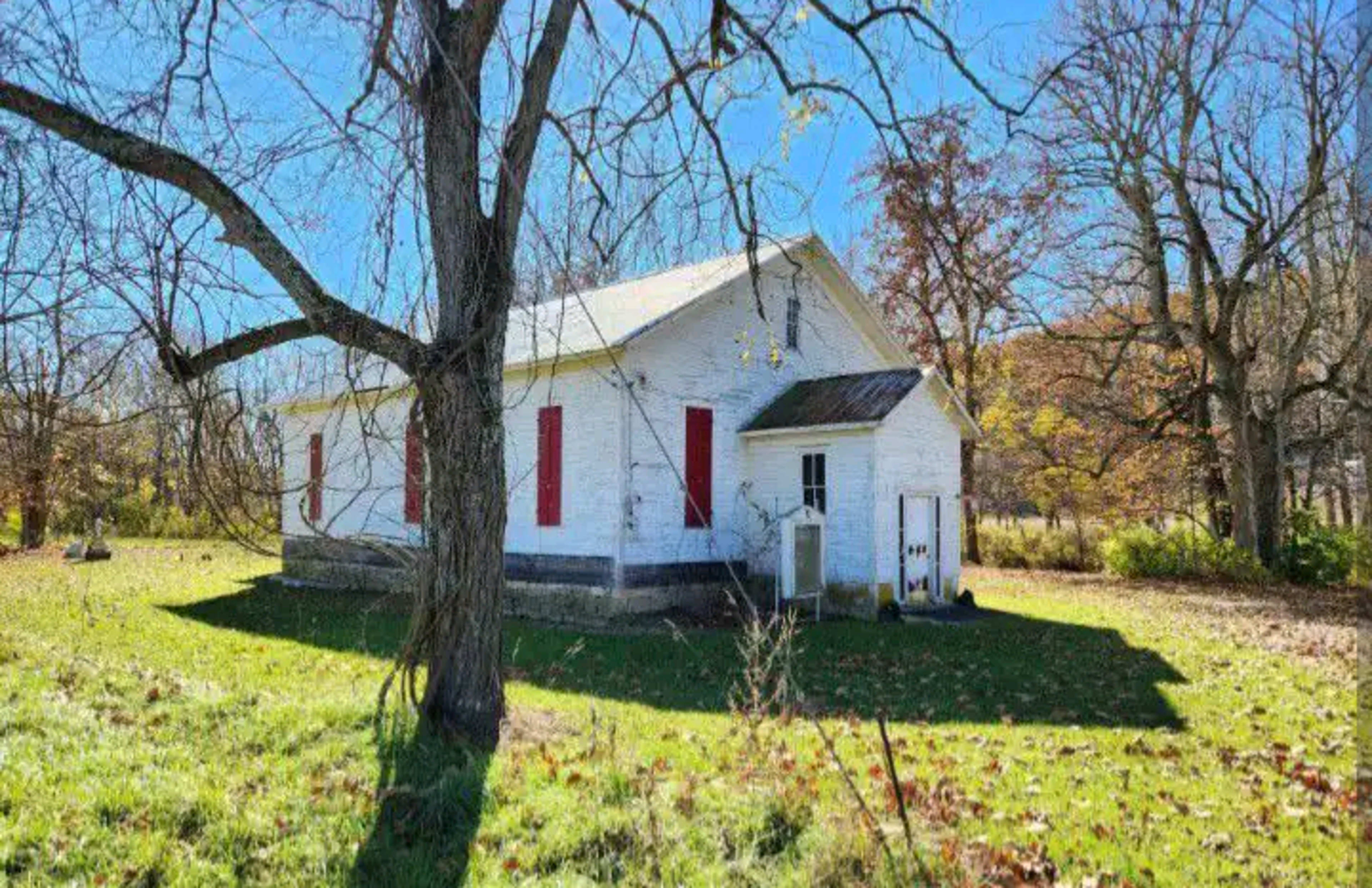 A white church with red shutters stands surrounded by grass and bare trees on a clear day.