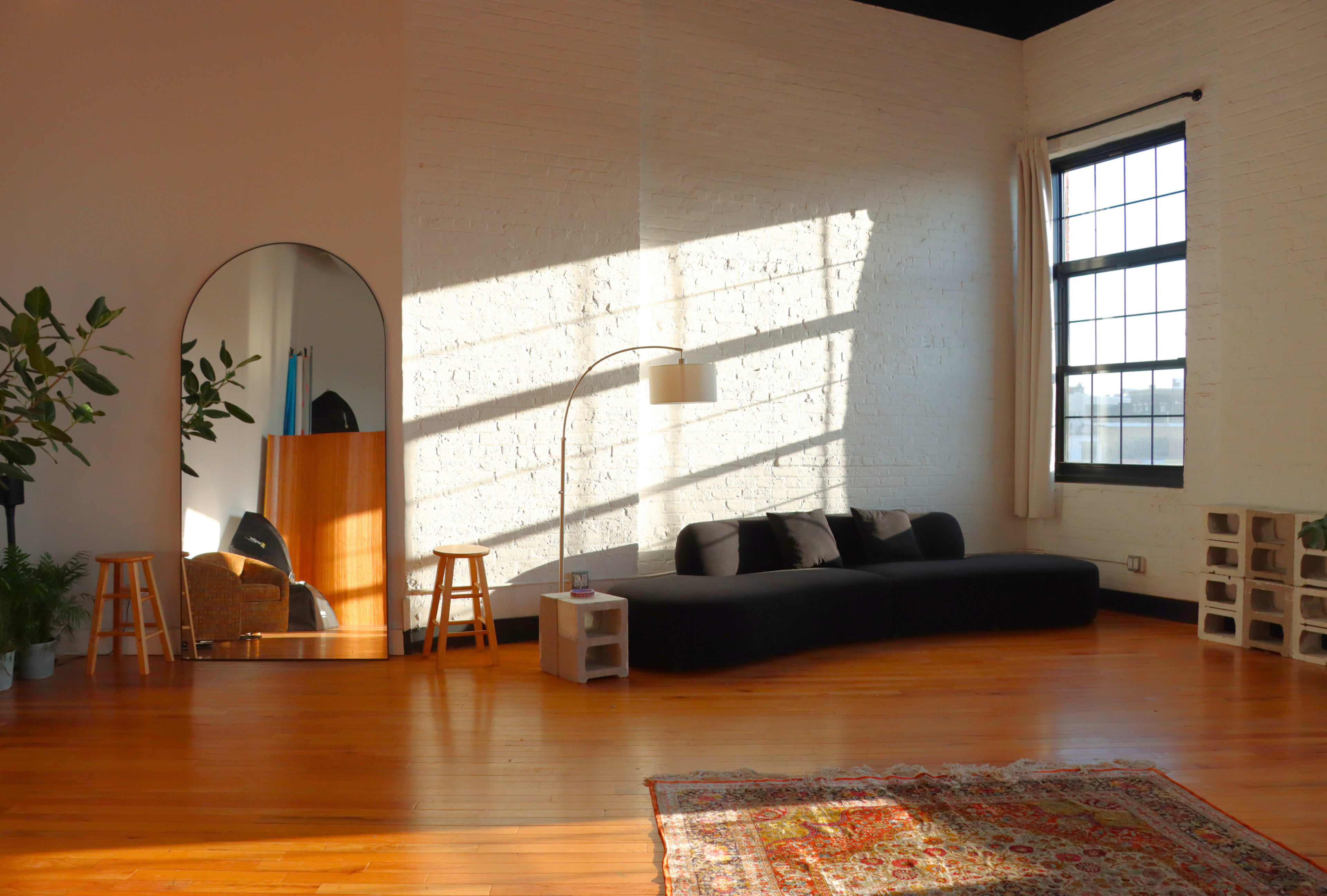 The image shows a spacious living room with a black curved sofa, a floor lamp, wooden stools, and a large mirror, illuminated by sunlight streaming through a window.