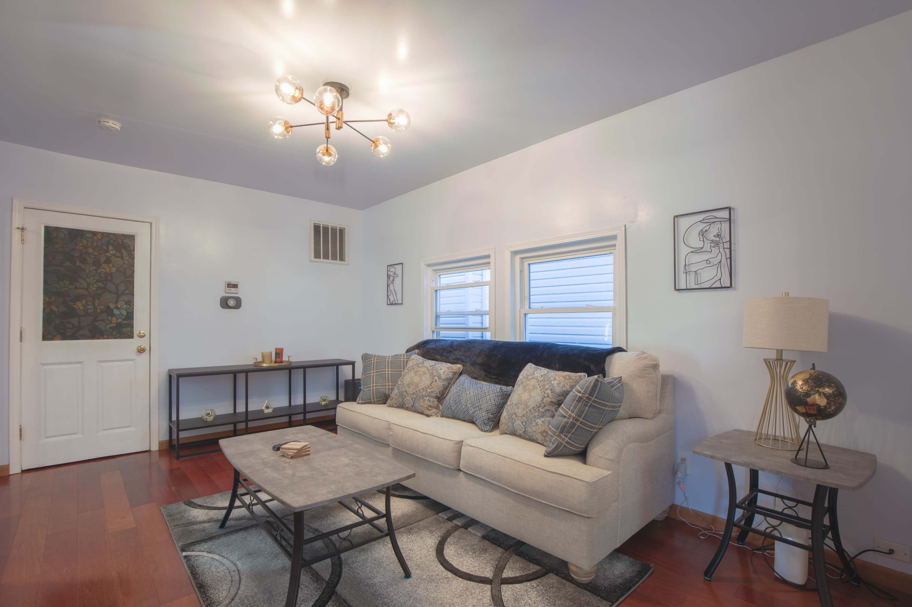 A modern living room features a light-colored sofa with patterned pillows, a round coffee table, and a lamp beside a decorative globe, all set against a wall with two windows and a door.