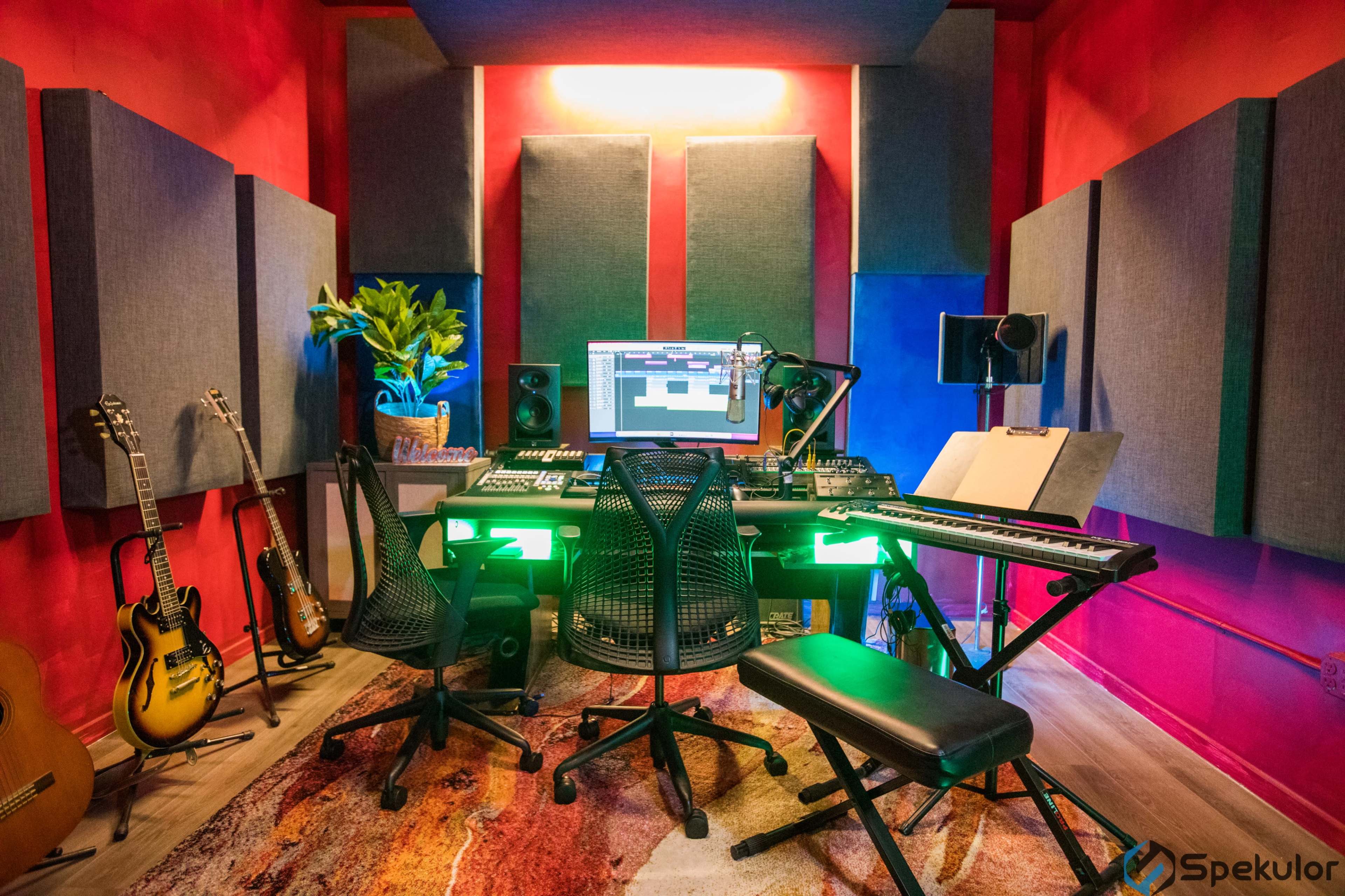 A music production studio features a central desk with a computer setup, surrounded by soundproofing panels, instruments on the walls, and a colorful, illuminated atmosphere.