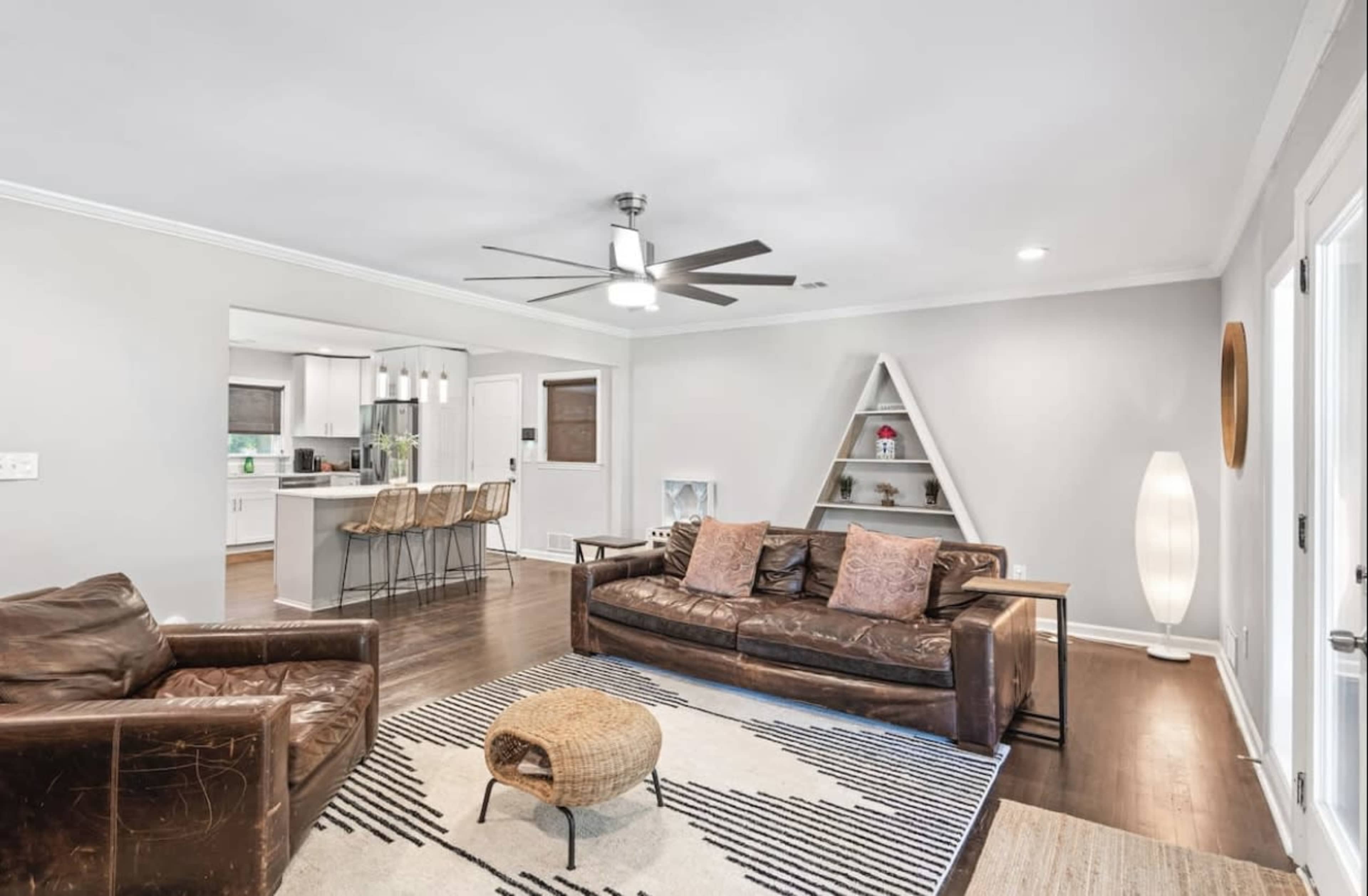 A living room features a brown leather sofa and chair with a round ottoman, a light-colored rug, and an open view into a kitchen area with bar stools.