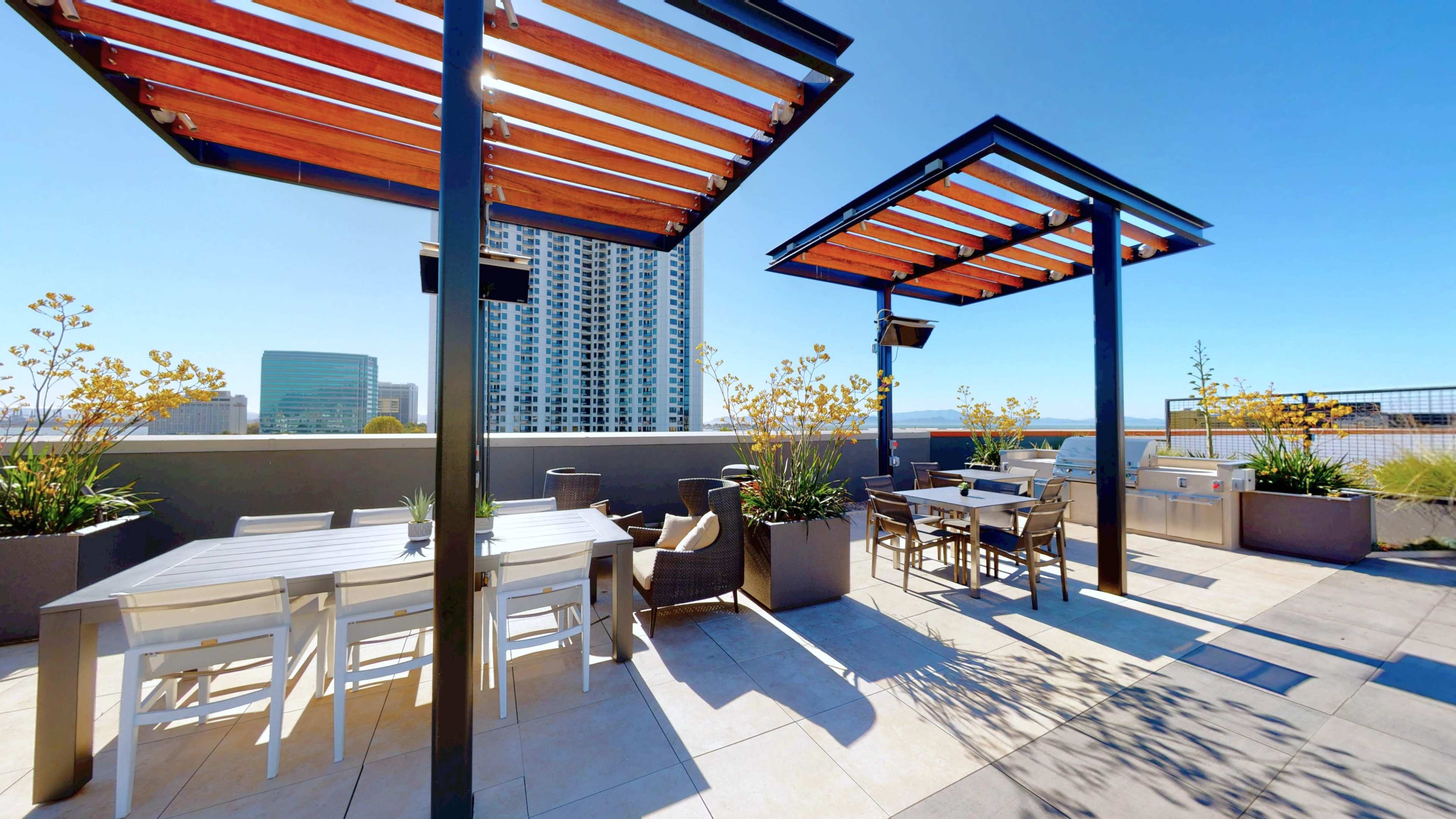 The image shows a rooftop terrace featuring seating areas, shaded pergolas, and planters with plants, alongside a city skyline in the background.