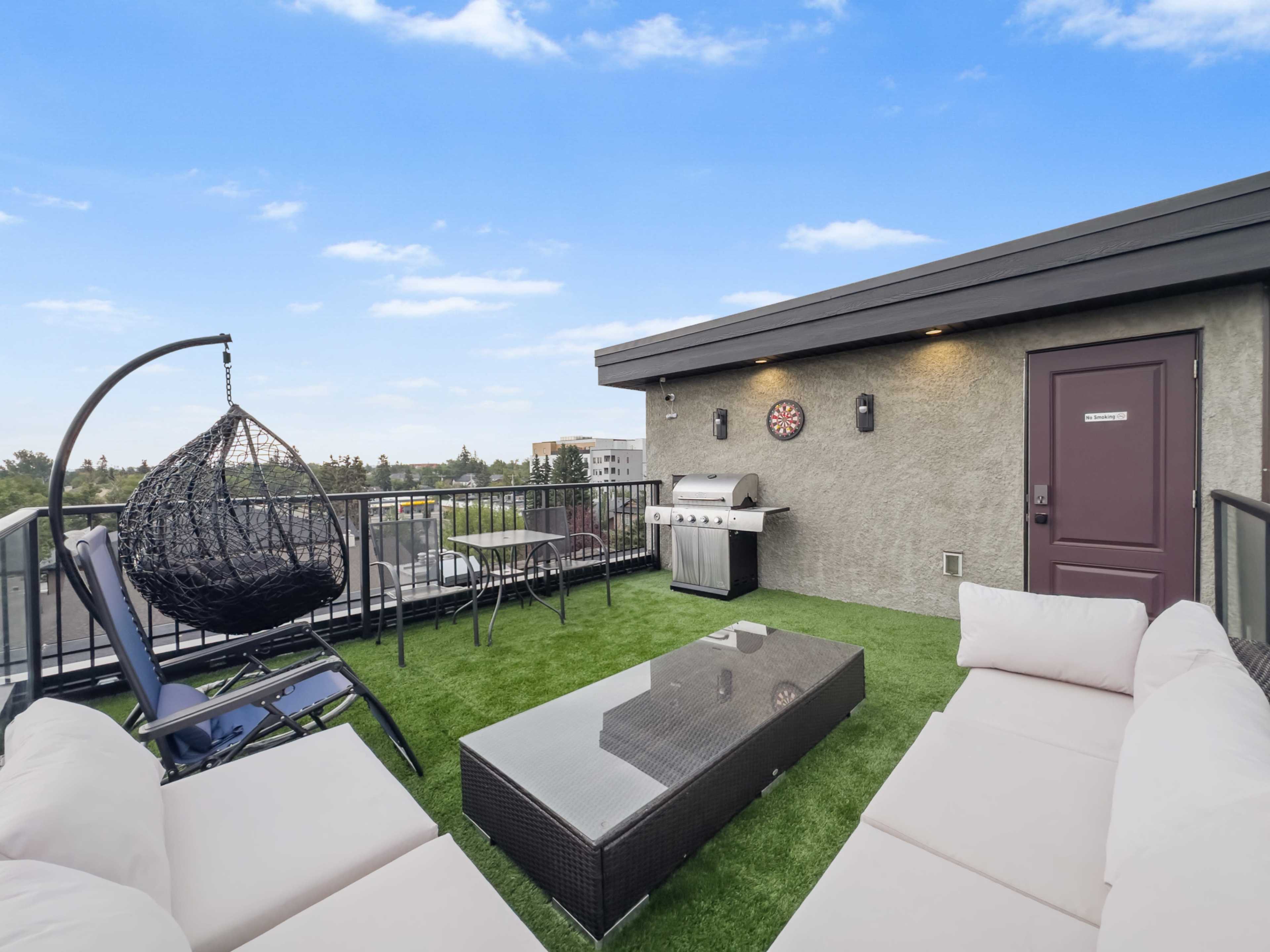 The rooftop space features a sectional sofa, a hanging chair, a grill, and a glass table on artificial grass with a view of the sky.
