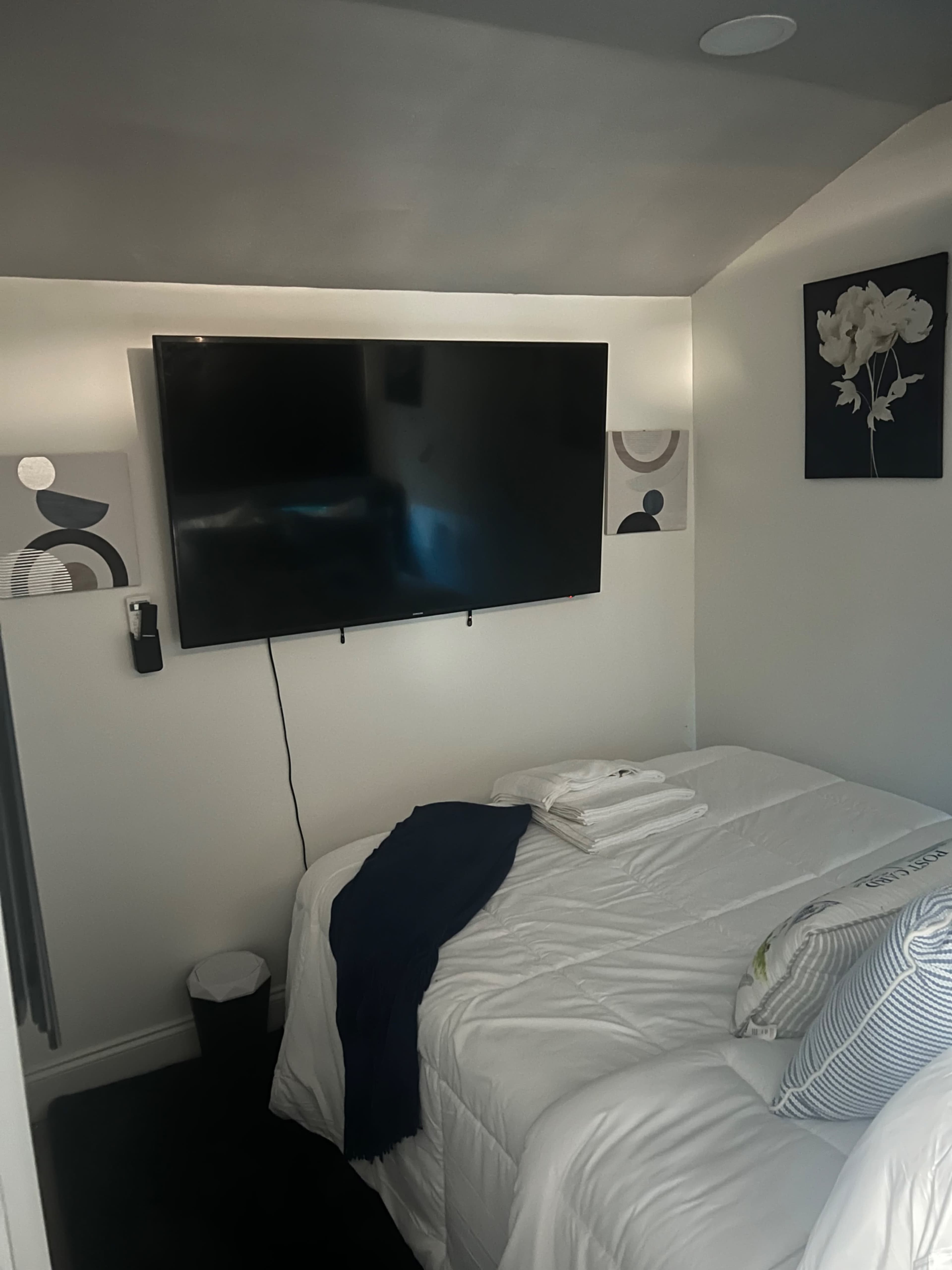 A small bedroom with a bed, a large wall-mounted TV, and minimal decorative art on the walls.
