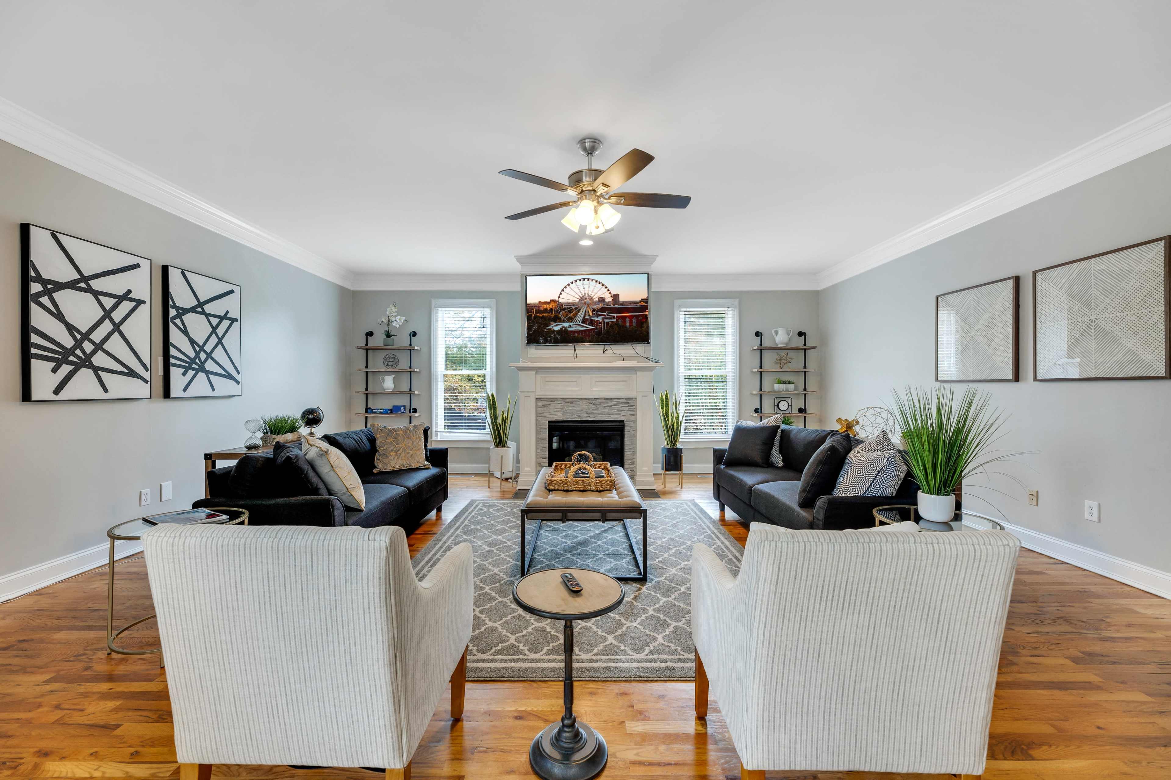 A spacious living room features two black sofas, two light-colored armchairs, a coffee table, and a fireplace, with decorative artwork on the walls and plants scattered throughout.