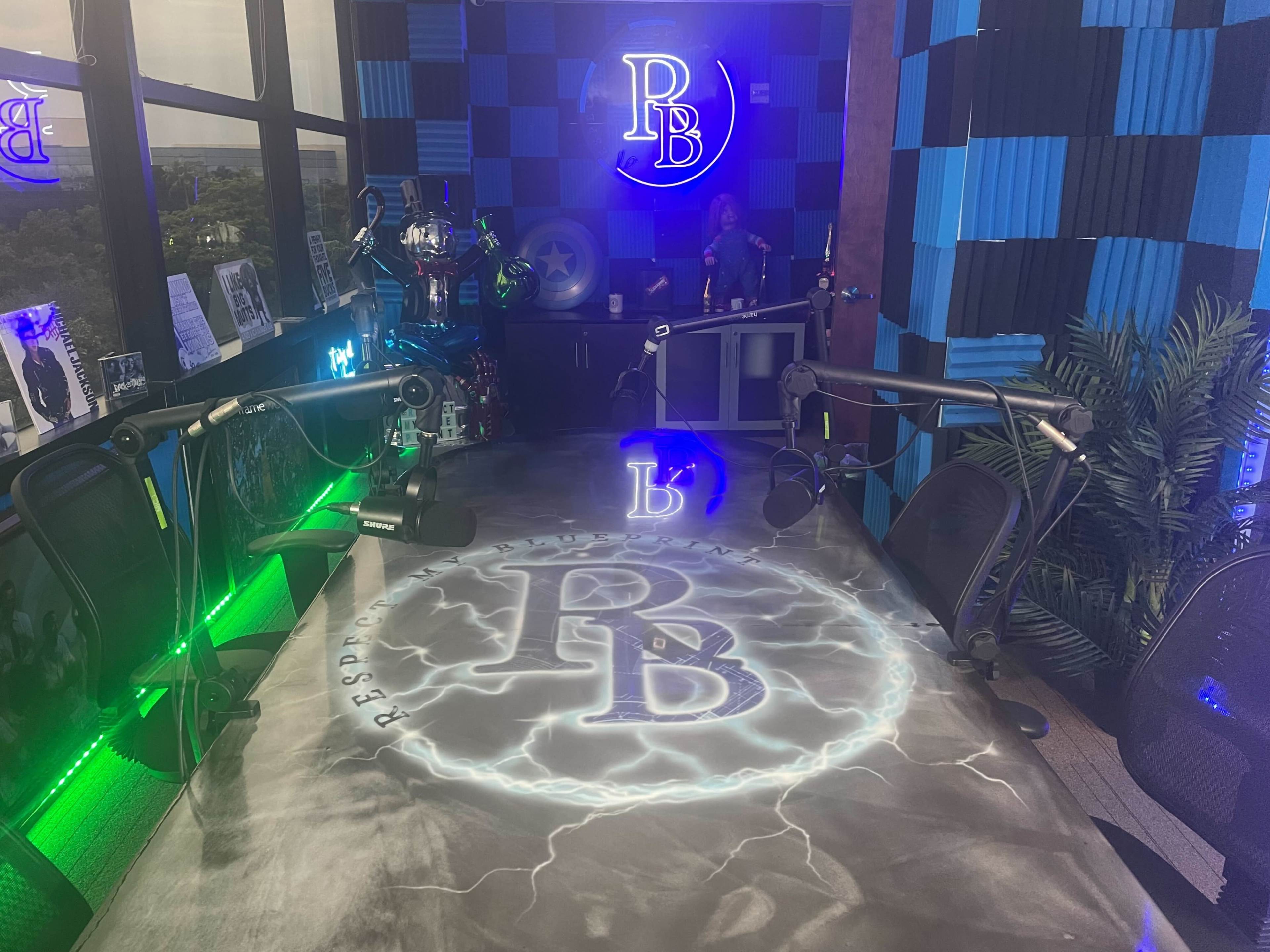 A brightly lit recording studio with a large table featuring the letters "PB" in a lightning design, surrounded by microphone stands and colorful acoustic paneling.