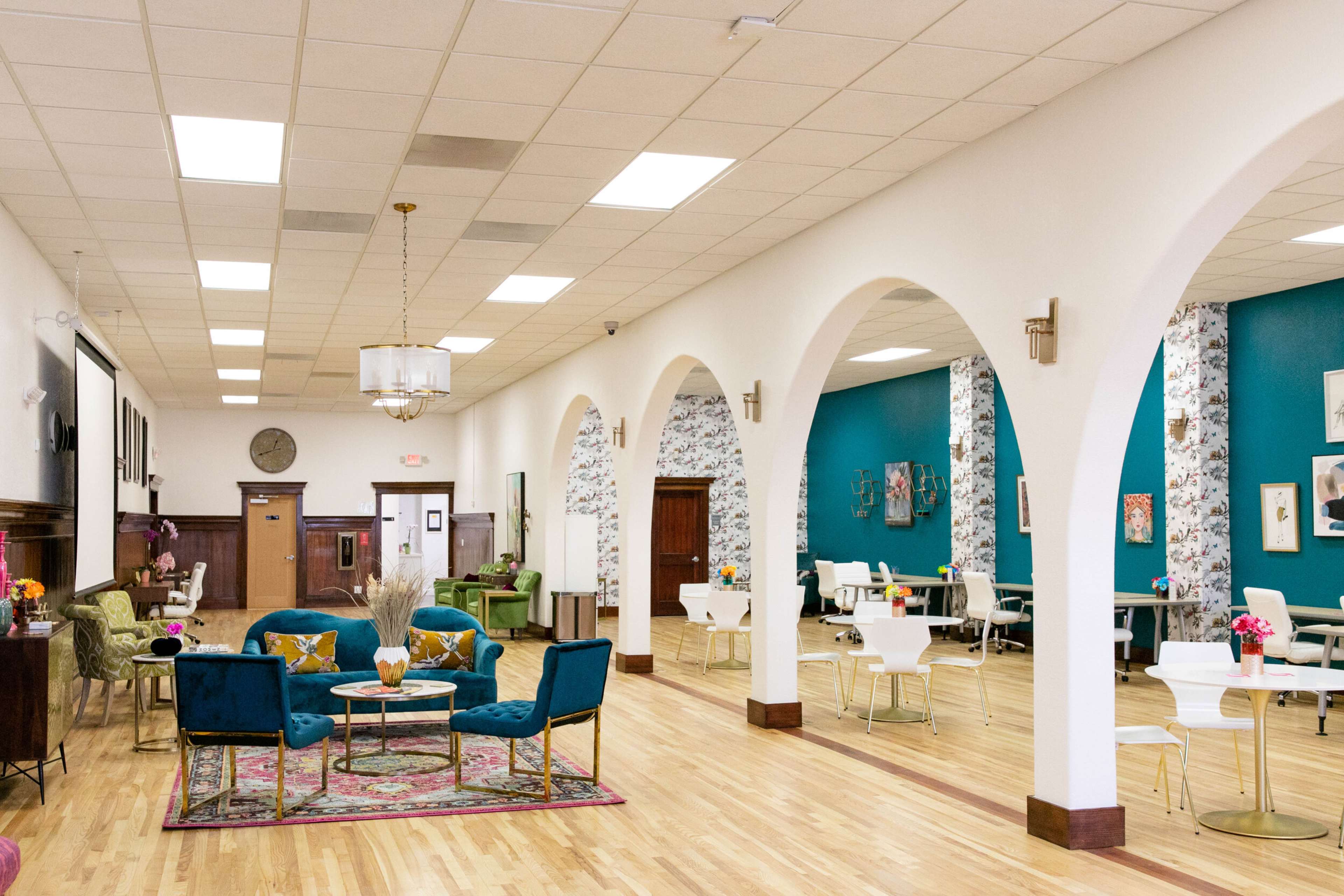 A spacious interior with archways, featuring seating areas with blue velvet chairs and tables arranged for dining or meetings against a backdrop of white walls and teal accents.