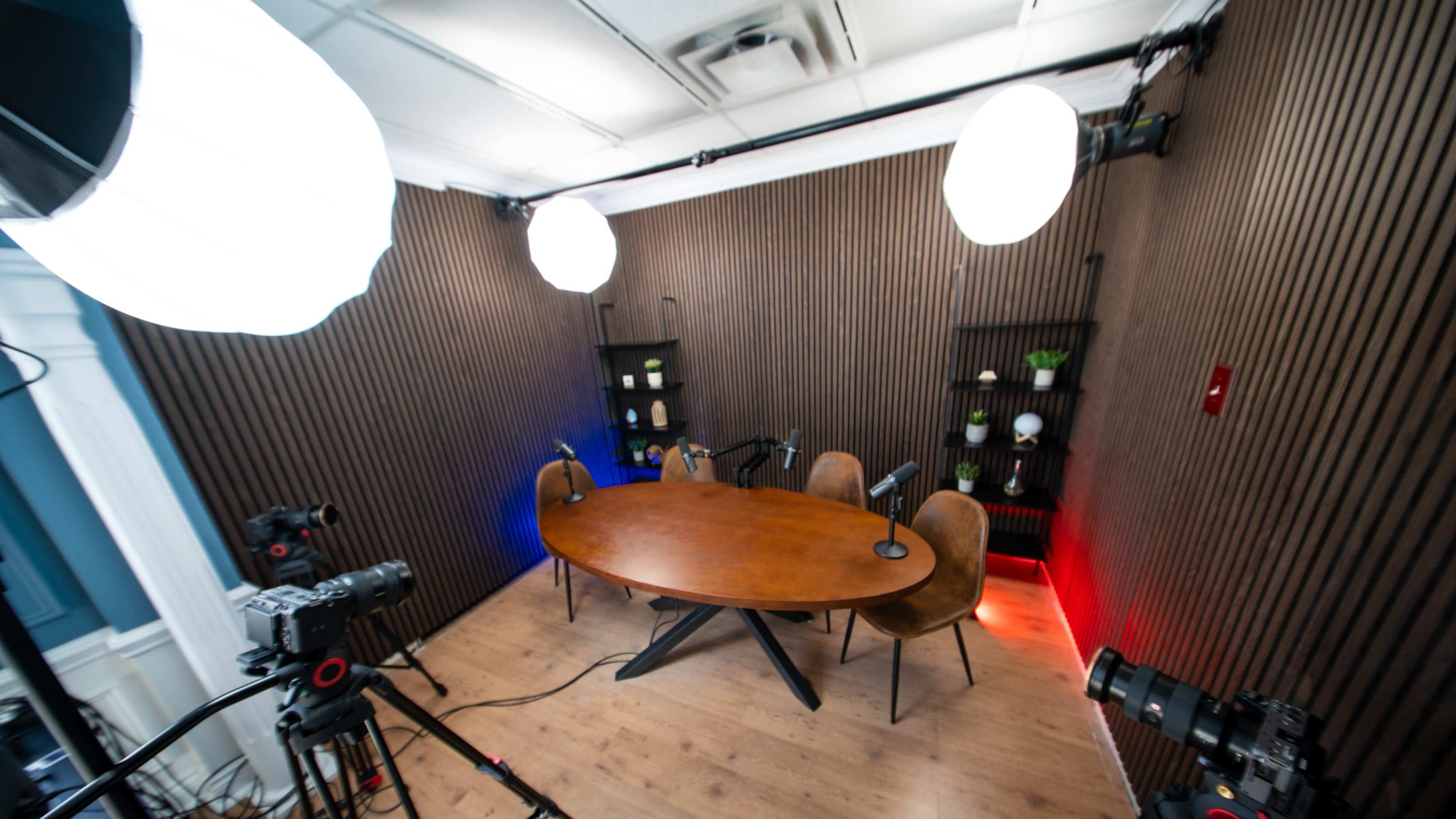 The image shows a podcast studio featuring a large wooden table surrounded by three chairs, with microphones set up and studio lights positioned nearby.