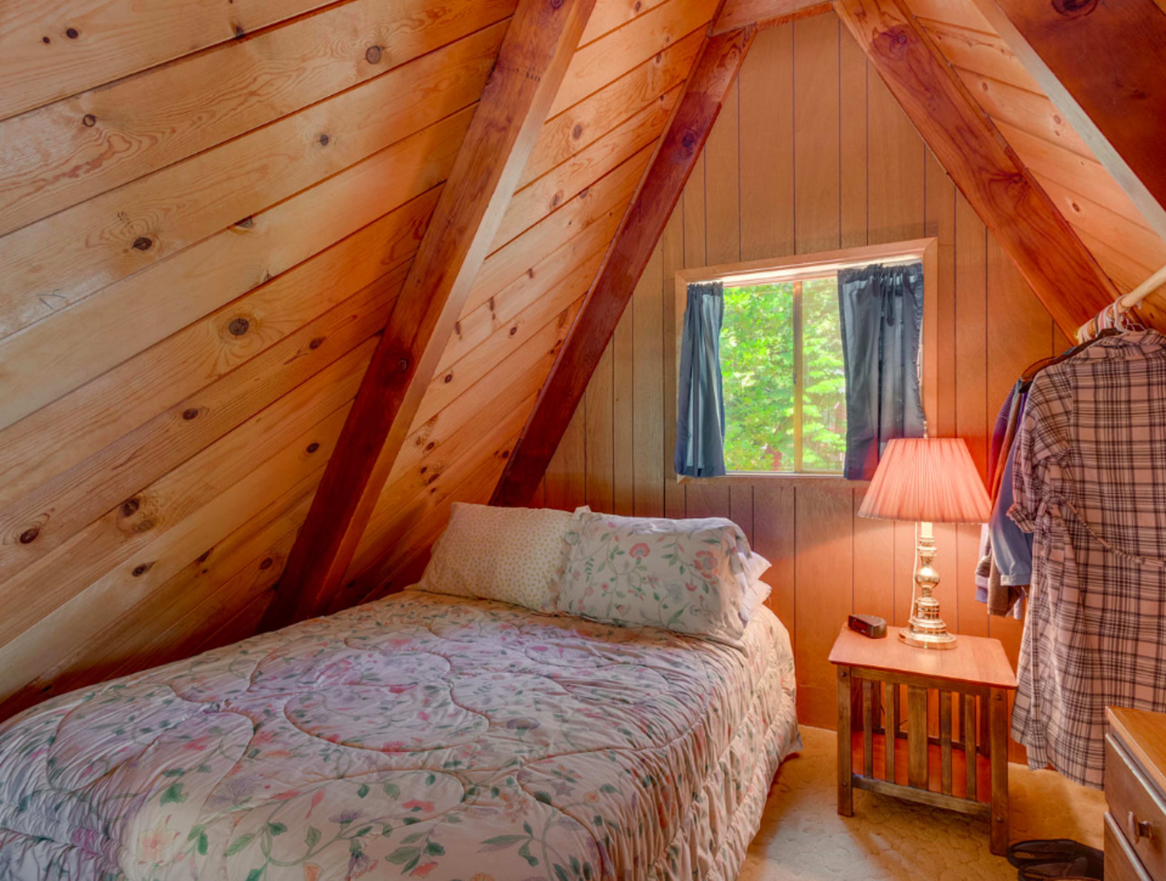 A small bedroom features an A-frame design with wooden walls, a single bed adorned with floral bedding, and a window with blue curtains.